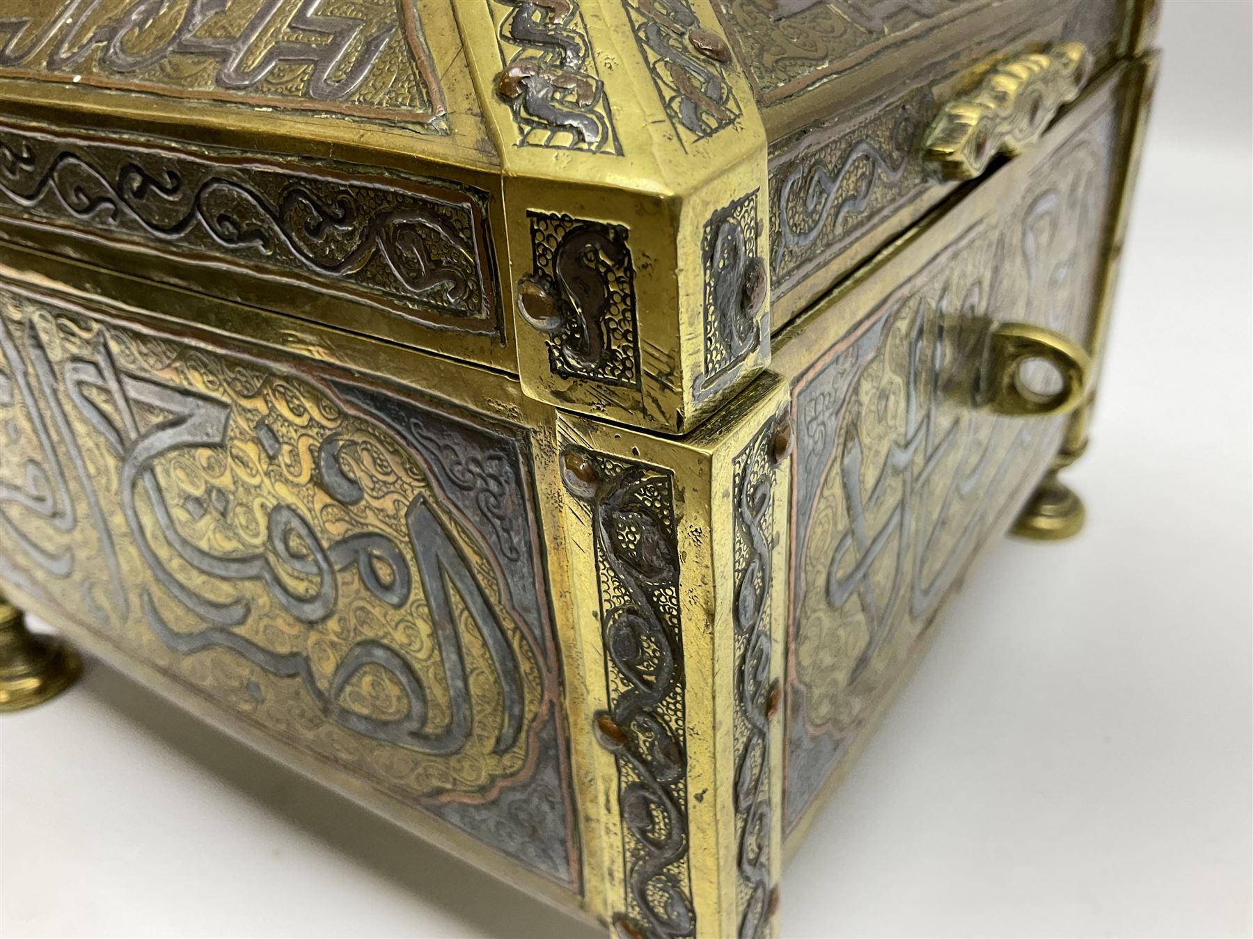 Late 19th century Damascus Cairoware Quran box, of square form with canted hinged cover, upon four circular pad feet, the brass exterior inlaid with silver and copper script and decorative detail, opening to reveal a marquetry lined interior, H15.5cm W19cm D19cm