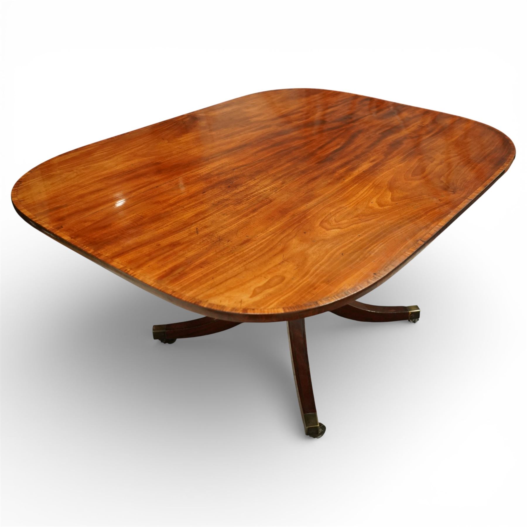 Regency figured mahogany breakfast table, figured rectangular tilt-top with rounded corners and rosewood banding, on turned column with four out-splayed supports with brass cups and castors 