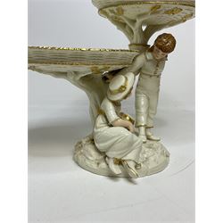 Royal Worcester figural double comport in the style of Kate Greenaway, modelled with a young boy and girl with painted features beside an oak tree stump branching out to support the two basket woven style dishes, raised upon rocky circular base, decorated with gilt detail throughout, with green printed stamp mark and impressed marks beneath, H19.5cm