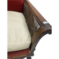 Early 20th century bergère three seat settee, shaped shell carved cresting rail, double caned back and sides, scrolled upright arms and shaped apron carved with shell and foliage, on hairy paw carved feet