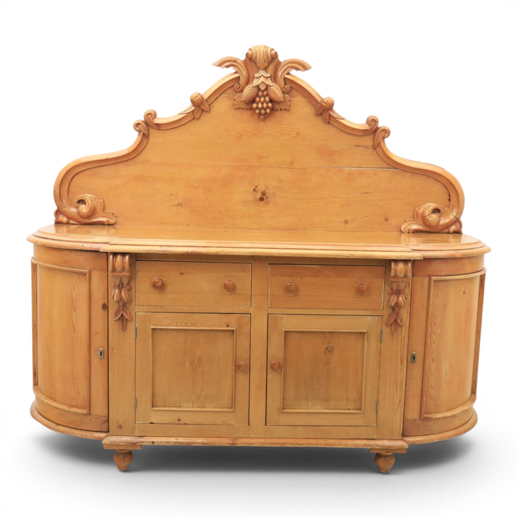 Victorian pine chiffonier, shaped moulded top with raised scroll carved back surmounted by carved fruit and foliate crest, fitted with two frieze drawers over two panelled cupboard doors, the outer cupboards of bowed form, carved foliate corbels, on plinth base