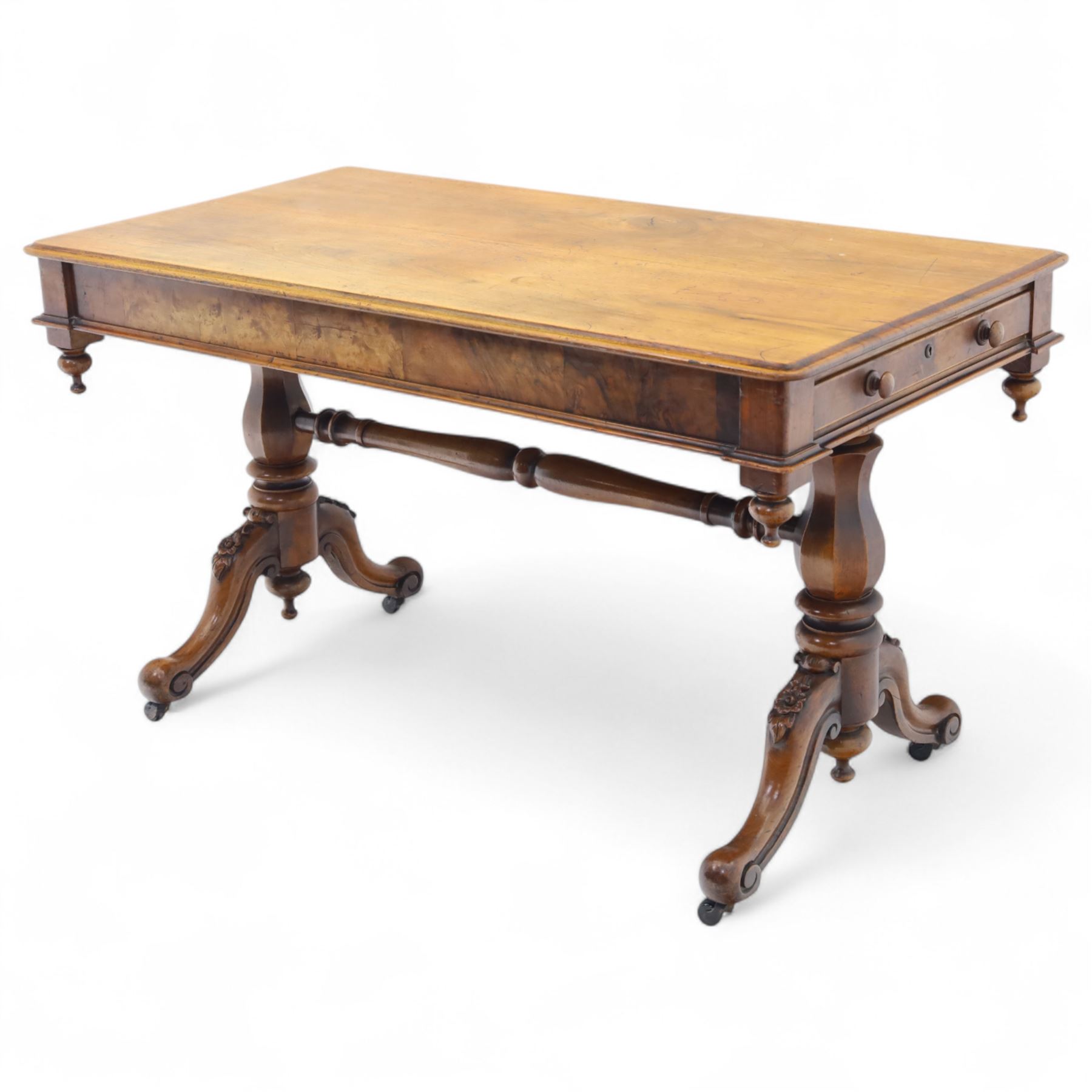 Mid Victorian walnut library table, plain top of rectangular form with moulded edge above frieze fitted with opposing drawers, table supported on a pair of turned and hexagonal carved baluster end standards, united by a turned stretcher terminating in scrolling sabre legs carved with acanthus and floral details, resting on ceramic castors, circa 1860