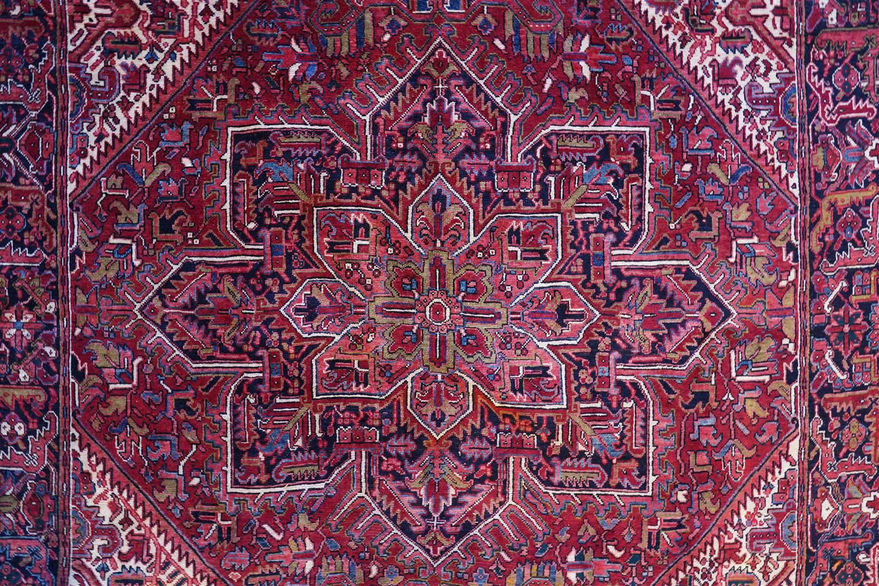 North West Persian Heriz hand-knotted carpet, brick-red field with a large central angular star medallion with pendant motifs, surrounded by stylized geometric palmettes, serrated leaves and tribal devices, ivory corner spandrels similarly decorated, field enclosed by a dark indigo main border of stylized rosettes and angular vine motifs between multiple guard stripes, with cotton fringe ends  