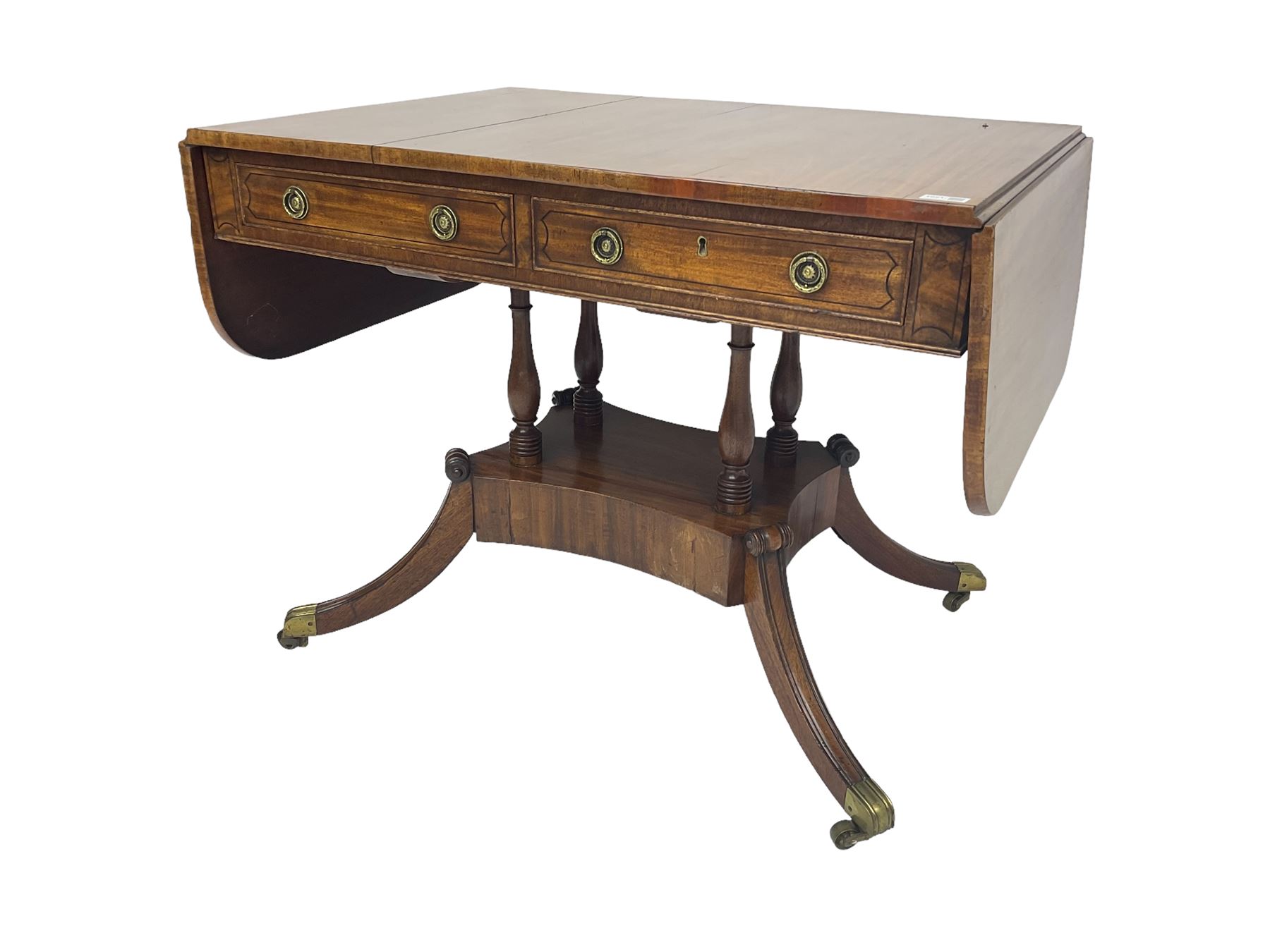 Regency period mahogany drop-leaf sofa table, rectangular top fitted with two cockbeaded drawers with ebony stringing, raised on turned columns united by quadrupod base, the sabre supports terminating in brass cups and castors