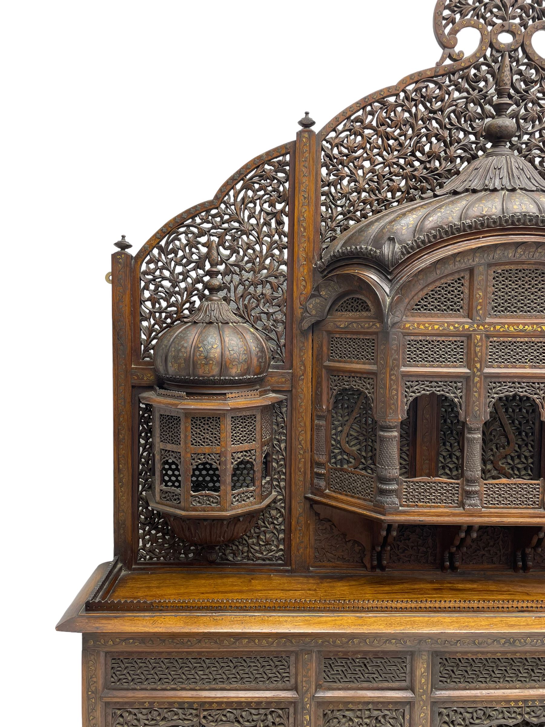 Late 19th century Indian Burmese carved and pierced hardwood and brass inlaid wall shelf, the shaped back profusely decorated with trailing and interlaced foliage, with projecting architectural structures, the central balcony with canopy top and five open windows with arched apertures on columns, two flanking five-sided balconies, the shelf supported by six foliate carved columns with pierced cusped pointed arches 