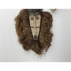 Two African carved masks, comprising of a white painted mask with carved facial details and a long form mask with carved eyes and open mouth surrounded with raffia decoration, longest example L60cm