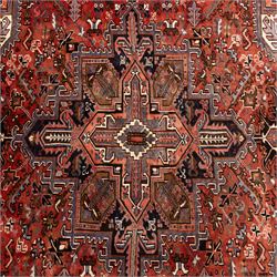 North West Persian Heriz hand-knotted carpet, madder red field with central stepped geometric medallion with radiating angular motifs and serrated outlines, and stylized floral and tribal devices framed by a broad border of repeating angular palmettes and rosettes flanked by multiple guard stripes, with cotton fringe ends 