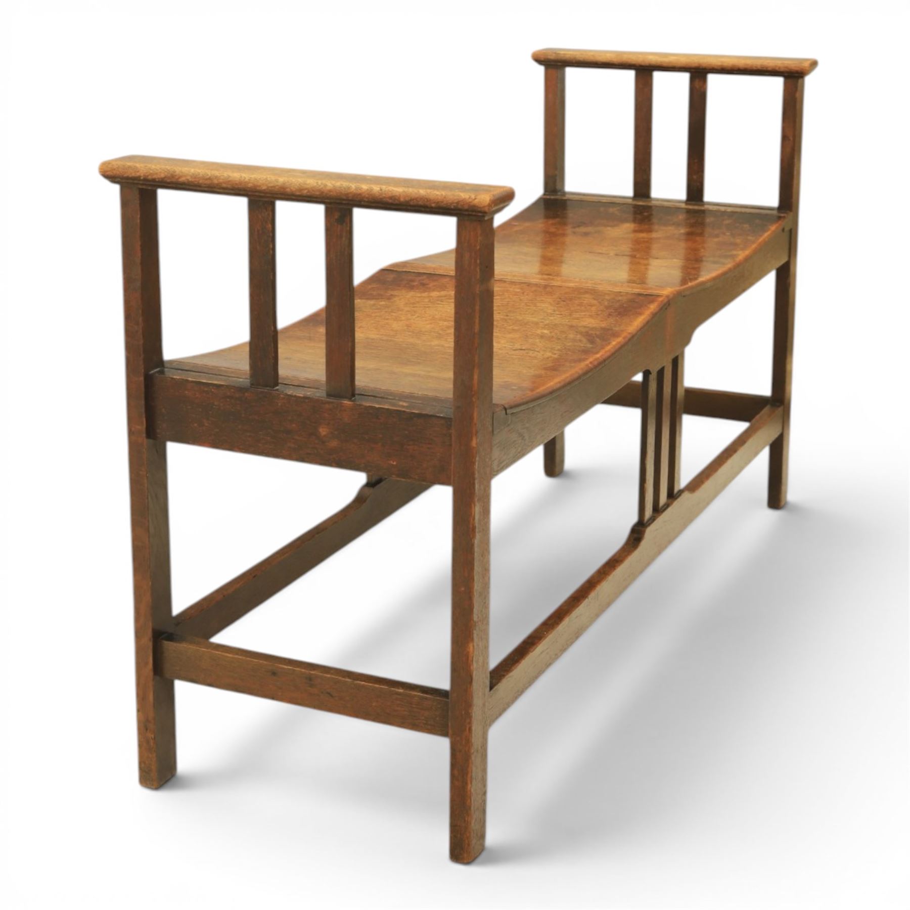 Arts and Crafts oak window bench, with flat armrests flanking two dished seats, raised on square supports united by stretchers