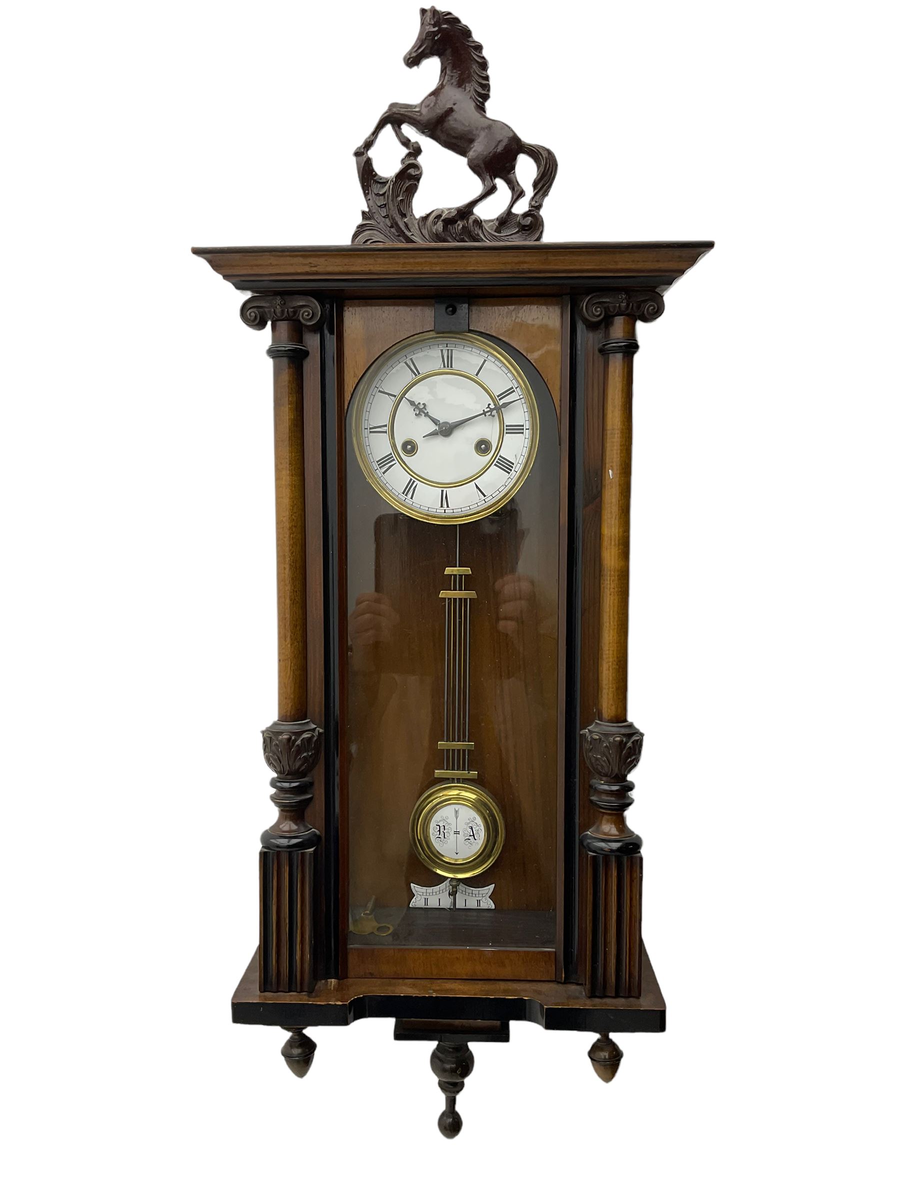 A late 19th century German spring driven 'Vienna' wall clock with a fully glazed door flanked by turned columns with ionic capitals and carving, replacement pediment depicting a prancing horse, with a two-piece enamel dial, Roman numerals, minute track and gothic steel hands, 8-day movement striking the hours and half hours on a gong,  with a gridiron pendulum and beat plate. With key.


