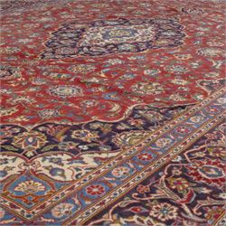 Central Persian Kashan hand-knotted carpet, madder red field with a central cusped medallion of ivory and deep indigo ground, enclosing a stylized floral rosette, framed by scrolling arabesques, field richly ornamented with an intricate network of flowering vinery, palmettes, and delicate floral sprays, spandrels in deep indigo, principal border of navy ground decorated with a continuous palmette and vine-scroll, flanked by multiple guard stripes, with cotton fringe ends 