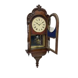 A 19thcentury American wall clock in a walnut case with crossbanding, with a carved pediment surround and arched glazed door flanked by two ring turned columns with pendants on an ogee plinth beneath, with a spring driven movement striking the hours on a bell, eight-inch painted dial with roman numerals, minute track and steel Maltese-Cross hands, with a spun brass bezel and winding collets, visible mock mercury pendulum with a reflective silvered mirror to the rear. With key and pendulum.   

