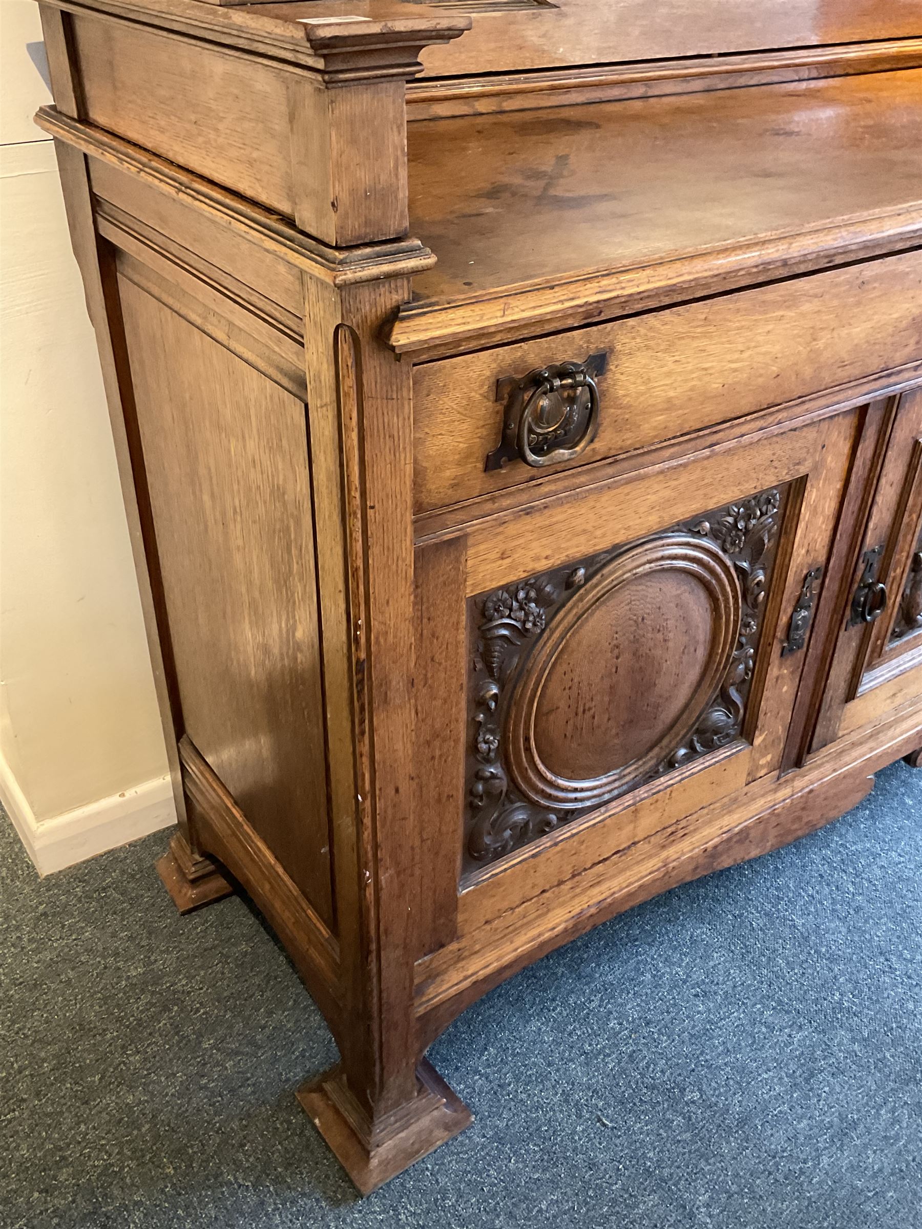Late 19th century Arts & Crafts period oak mirror back sideboard, shaped bevelled mirror back carved with foliate scrolls and flower heads, down swept supports with elongated club-shaped uprights, moulded rectangular top over two drawers and three cupboards, the drawer fronts with copper handles decorated with sailing ships, panelled doors carved with mythical beasts, flowerheads and foliage, on moulded block feet