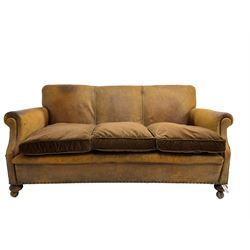Early-to-mid-20th century three seat sofa, upholstered in worn tan leather with studwork, loose cushions upholstered in brown fabric, on compressed bun feet with castors
