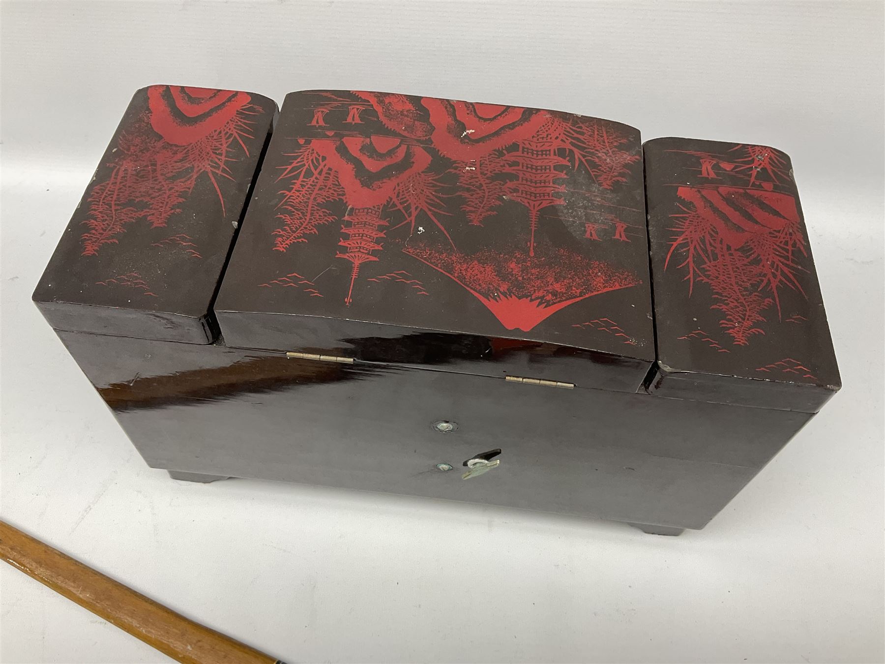 Oriental lacquered jewellery box, the body decorated with a black and red mountainous riverscape enclosing a glass compartment with simulated ivory rickshaw figure and three hinged lids opening to reveal mirror and various compartments and painted ceremonial stick