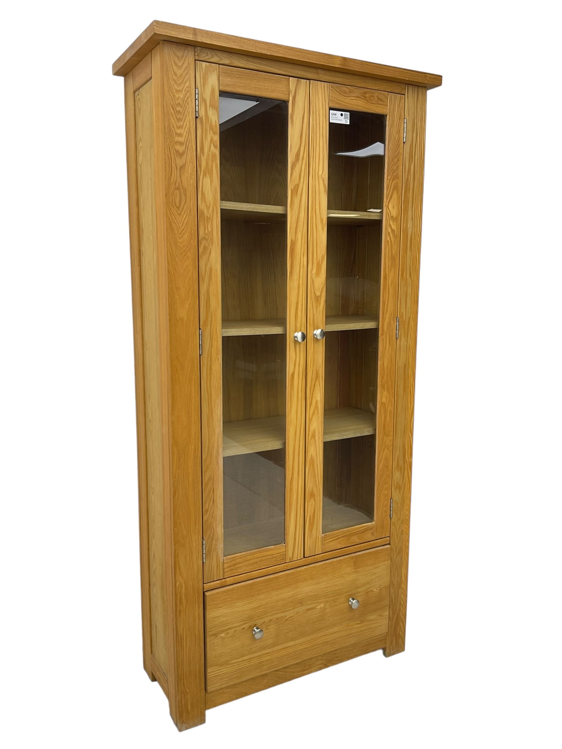 Light oak bookcase display cabinet, projecting square cornice over glazed double doors enclosing three adjustable shelves, single drawer base, on stile supports