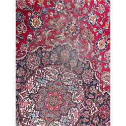 Large Persian Kashan crimson ground carpet, central floral medallion surrounded by swirling leafy branches and palmettes, enclose by floral pattern spandrels, the indigo border with overall scrolling design decorated with palmettes, within guard stripes 