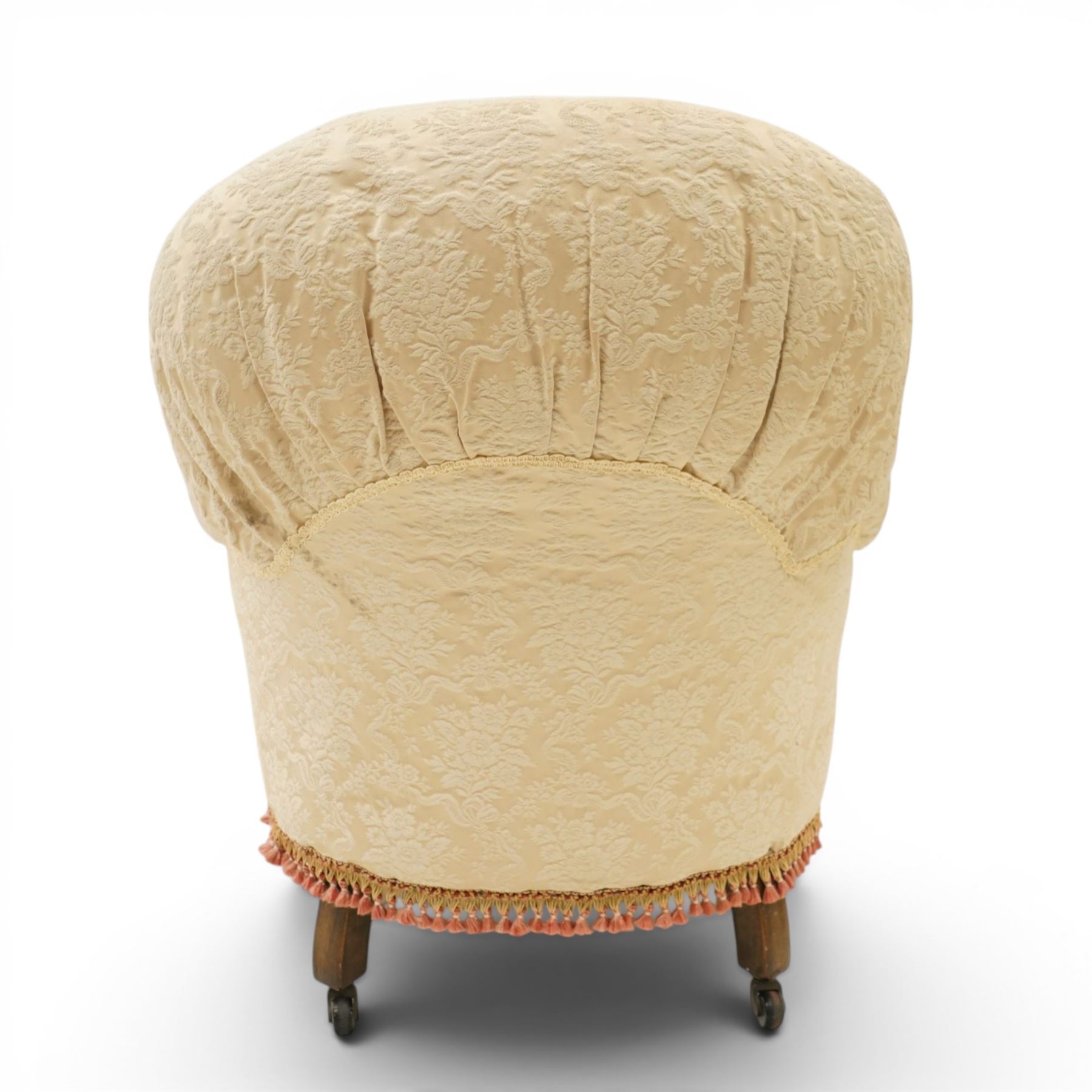 Victorian button-back nursing chair, the rounded horseshoe back enclosing a deep upholstered seat, padded arms and sprung seat in cream floral damask fabric, with crimson piping and tasselled lower fringe, on turned front legs with castors