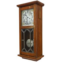 Art Deco style oak wall clock, moulded cornice above a silvered dial with Arabic numerals and geometric spandrels, glazed door with geometric bevelled glass panels enclosing a circular pendulum, on a stepped moulded base