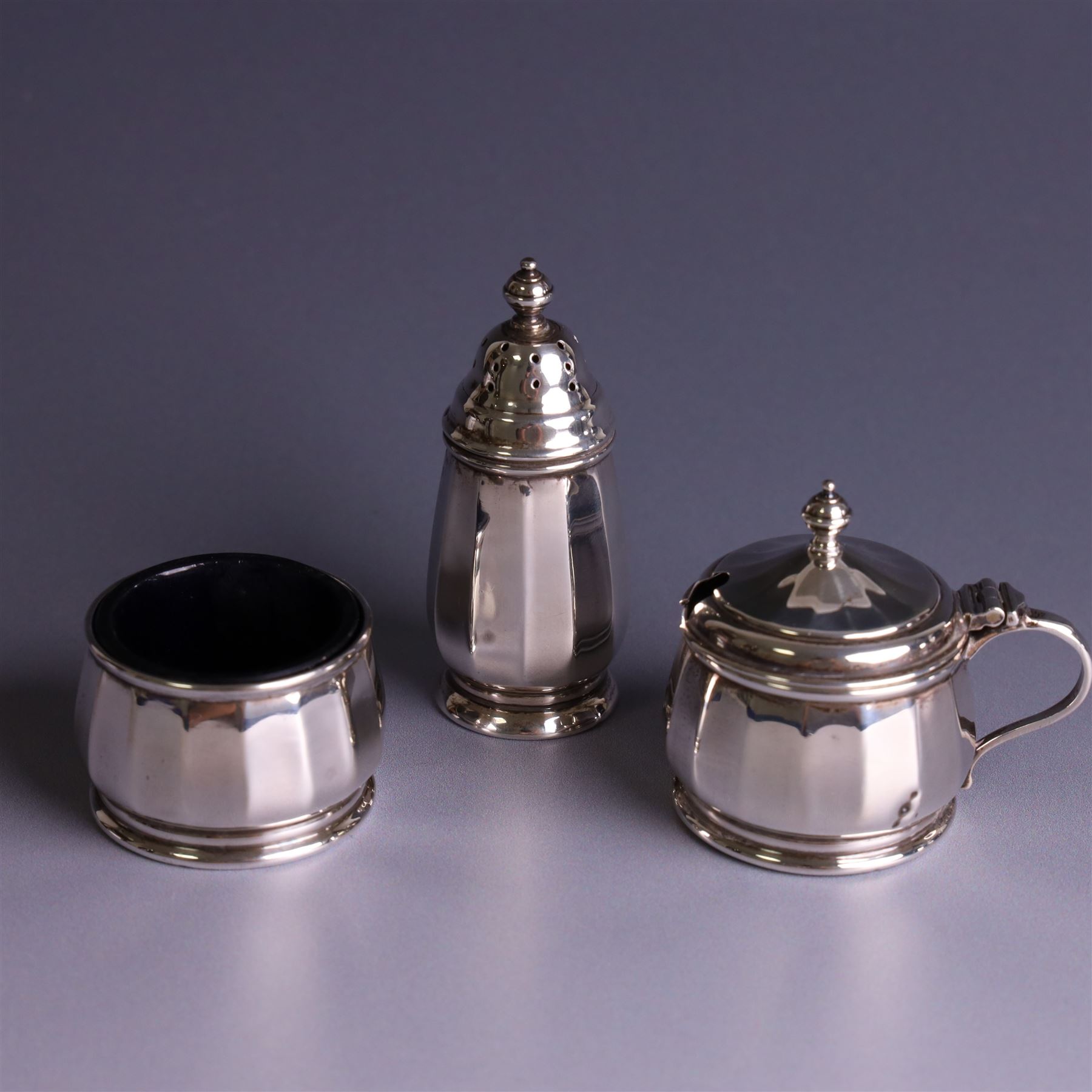 Mid 20th century three piece silver cruet set, comprising pepper shaker, open salt and mustard pot with cover, each of circular faceted form, hallmarked Walker & Hall, Birmingham 1958, the mustard pot and salt with blue glass liners