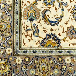 Central Persian Kashan hand-knotted carpet, ivory ground, with an all-over floral design of palmettes, scrolling vines and flowerheads in blue, ochre and beige tones, enclosed by a sage green border with repeating floral sprays and leafy scrolls, flanked by narrow guard stripes, with cotton fringes