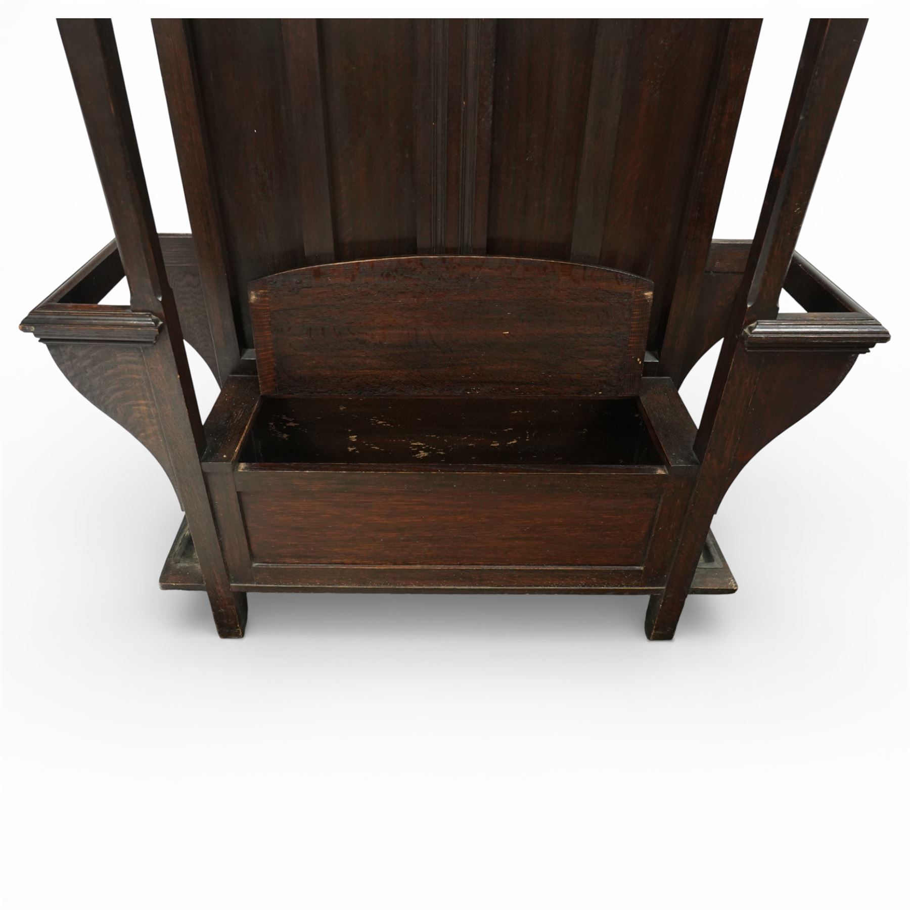 Arts and Crafts design oak settle, with projecting moulded cornice over a boarded back fitted with a rectangular bevelled mirror plate and a row of wrought iron coat hooks, the panelled back with arched fielded panels, flanked by shaped umbrella or stick stands with removable drip trays, the plank hinged seat over a panelled box base, on square section supports