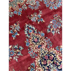 Persian Kirman crimson ground carpet, the field decorated with central floral medallion on a plain field decorated with flower head clusters, the outer field decorated with blue flower heads and stylised plant motifs, overall floral design border with trailing foliage branches, within guard bands 