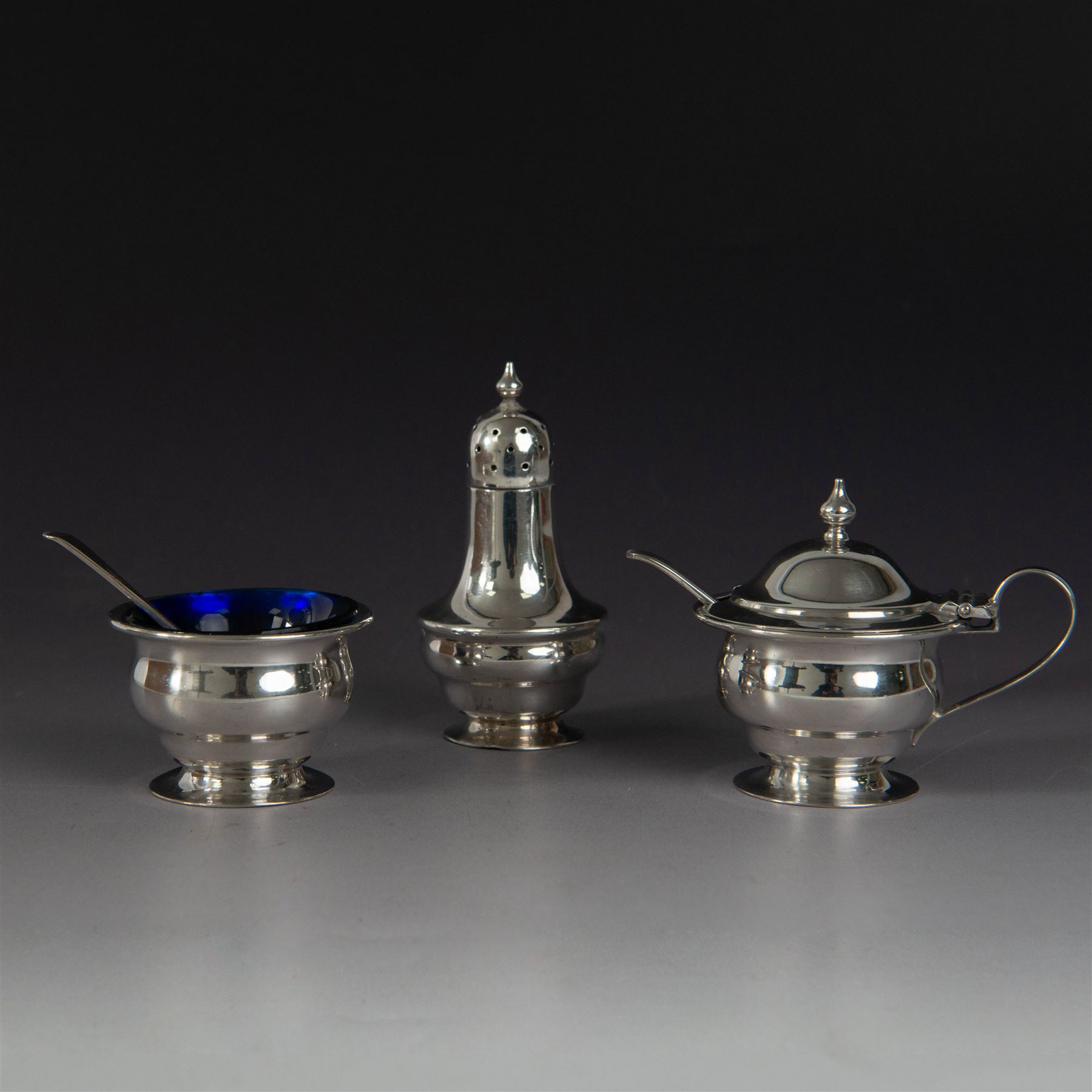 1930s silver three piece cruet set, comprising pepper shaker, open salt and mustard pot and cover, all of circular form upon circular spreading feet, hallmarked 	Atkin Brothers, Sheffield 1930, with two silver condiment spoons and blue glass liners