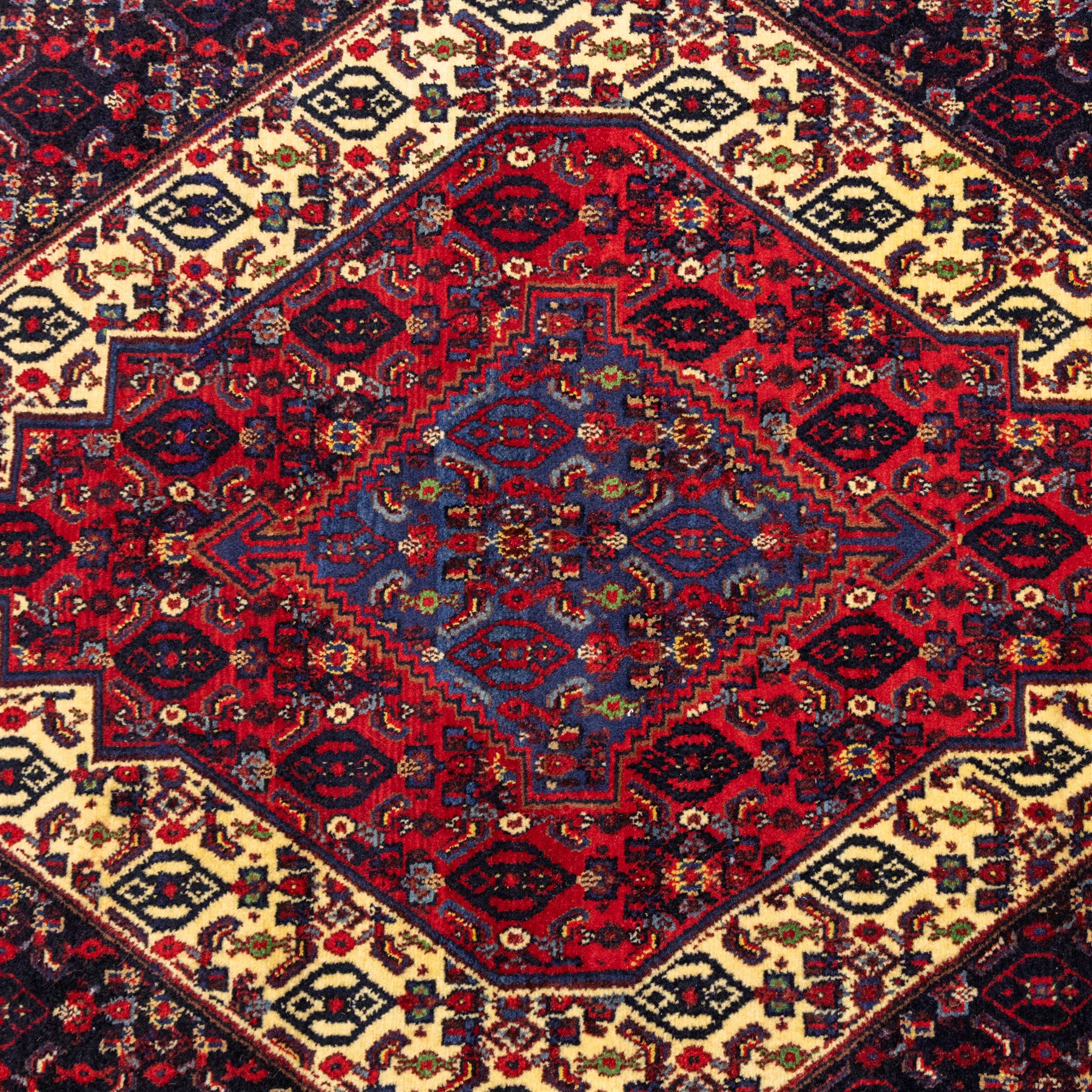 North West Persian Senneh hand-knotted rug, dark indigo field with a central bold stepped lozenge medallion of madder red and deep blue, enclosed within an ivory serrated contour forming a cruciform outline, medallion with repeating boteh and floral devices, field decorated with an all-over Herati pattern of rosettes enclosed by angular leaves principal border of madder red ground decorated with alternating floral heads, flanked by narrow guard stripes, with cotton fringe ends 