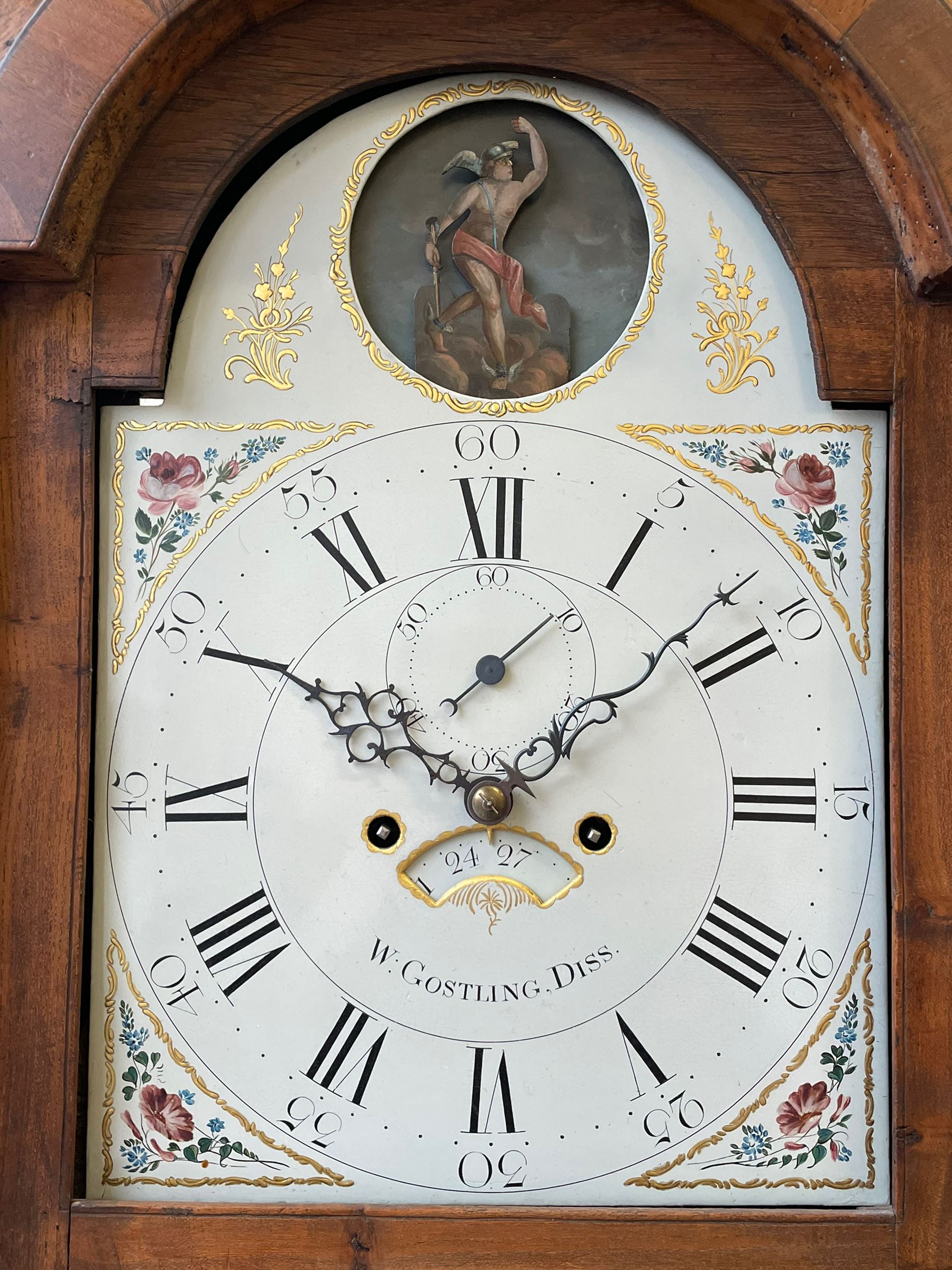 William Gostling of Diss  (Norfolk)-  Late 18th century 8-day oak longcase clock c1780-90, break arch top with scalloped cresting and three brass finials, glazed break arch hood door flanked by turned pilasters with brass capitals, trunk with a long trunk door and rounded top on a square plinth with an applied and shaped skirting, early painted Wilson of Birmingham dial with Roman numerals and five minute Arabic's, matching steel hands and second pointer, arched square ended calendar aperture with date disc behind, floral spandrels within gesso borders and a painted rocking automaton representing Mercury to the break arch, dial pinned via a false plate to a weight driven rack striking movement with a recoil anchor escapement, striking the hours on a cast bell. With pendulum weights and key. 
William Gostling (II) is recorded as working in Diss from 1774 to 1797, succeeded by widow Anne and son George . The dial of this clock is illustrated and discussed in detail in M.F. Tennants book 