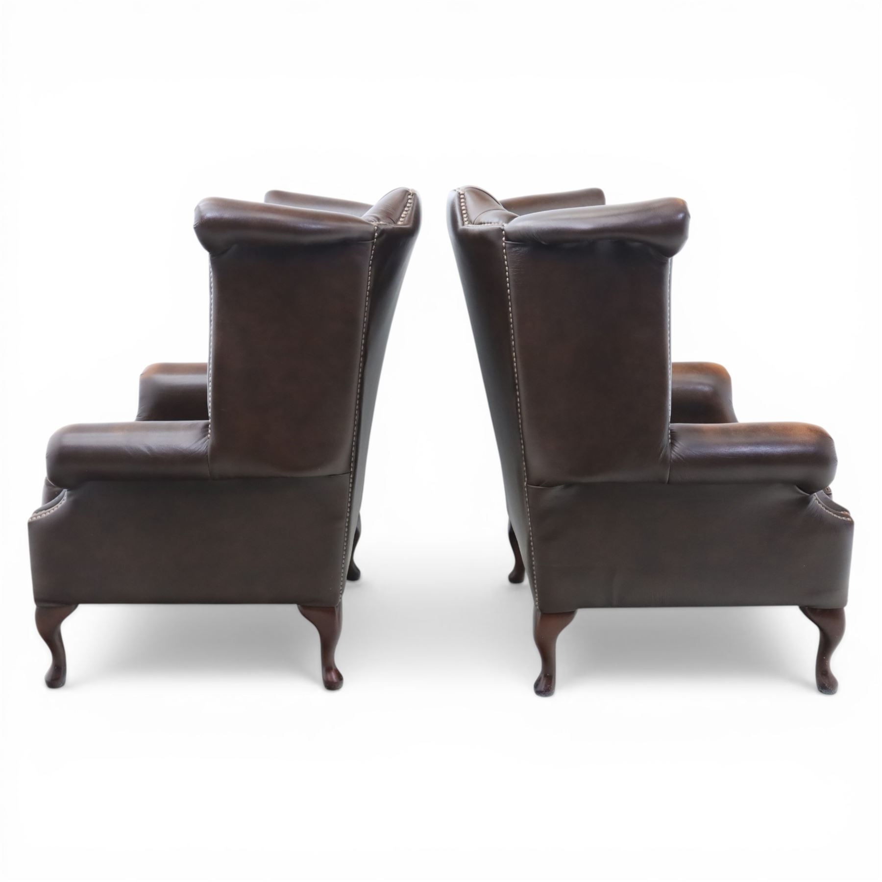20th century Chesterfield style brown leather wingback armchairs, with button upholstered backs and wings, shaped scroll arms with studded borders, loose seat cushions, raised on cabriole supports with pad feet