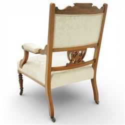 Edwardian mahogany open armchair, the upholstered rectangular back buttoned in ivory patterned fabric within a shaped top rail with inlaid foliate detail, open splat carved with a pierced anthemion motif centred by an inlaid roundel, swept open arms with upholstered pads, over a sprung upholstered seat, on turned front supports with brass castors