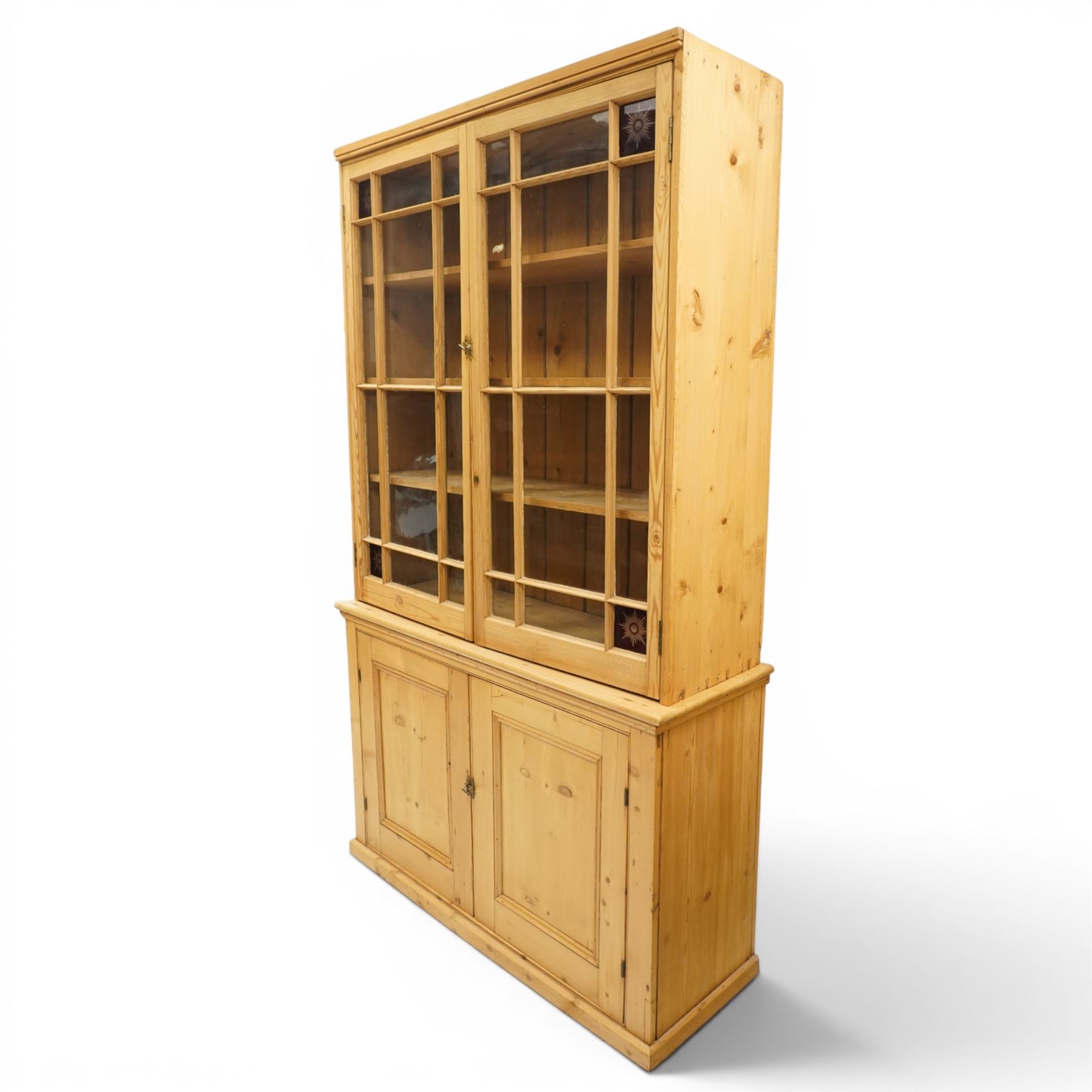 19th century pine glazed bookcase cupboard, twin astragal glazed doors with stained glass corner panels, enclosing fixed shelves, the lower section fitted with two panelled cupboard doors enclosing single shelf, on plinth base