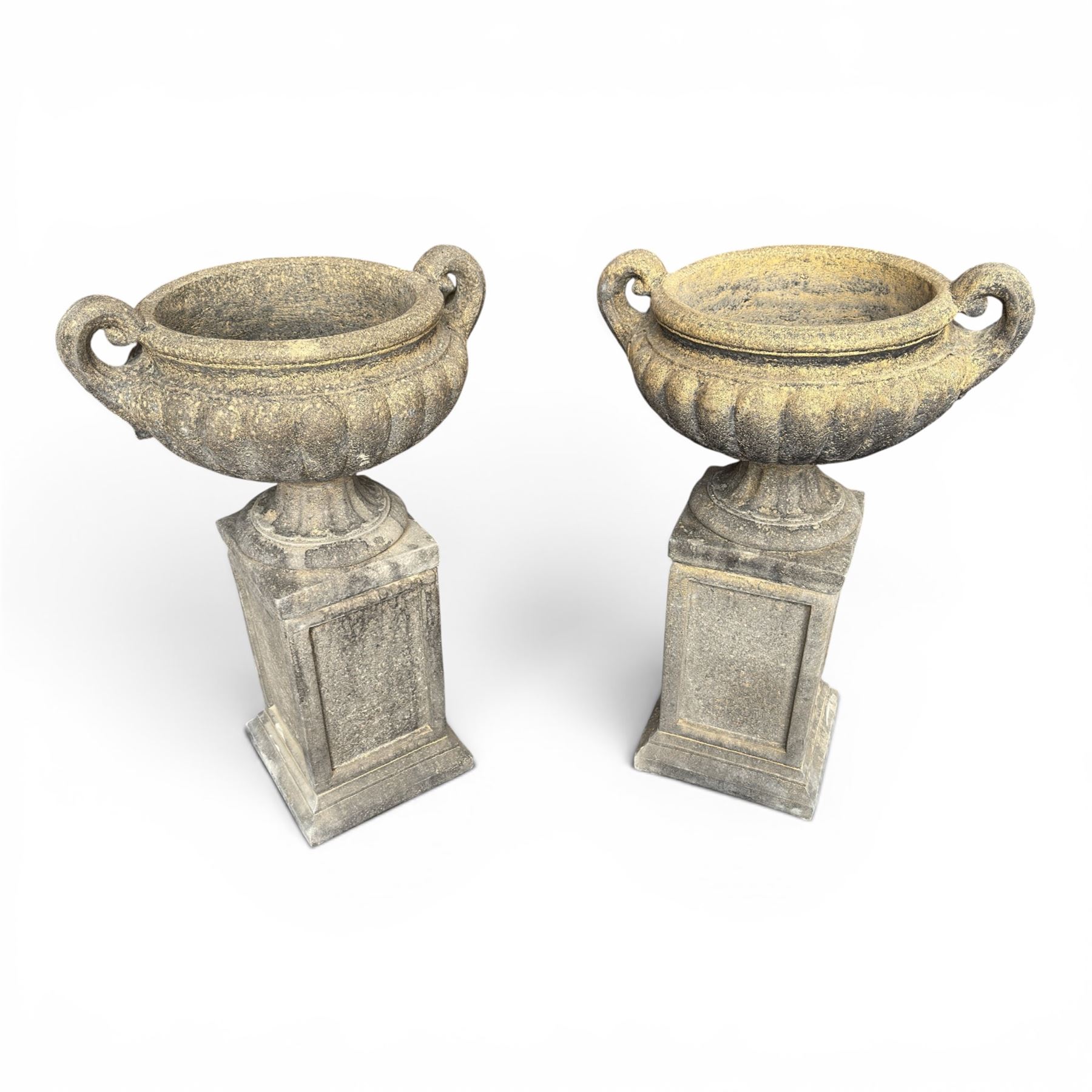 Pair of Neoclassical design weathered cast stone garden urns of kylix form, the rolled rim flanked by twin pierced scroll handles over a shallow gadrooned bowl, raised on a fluted circular socle and faux panelled square pedestal base