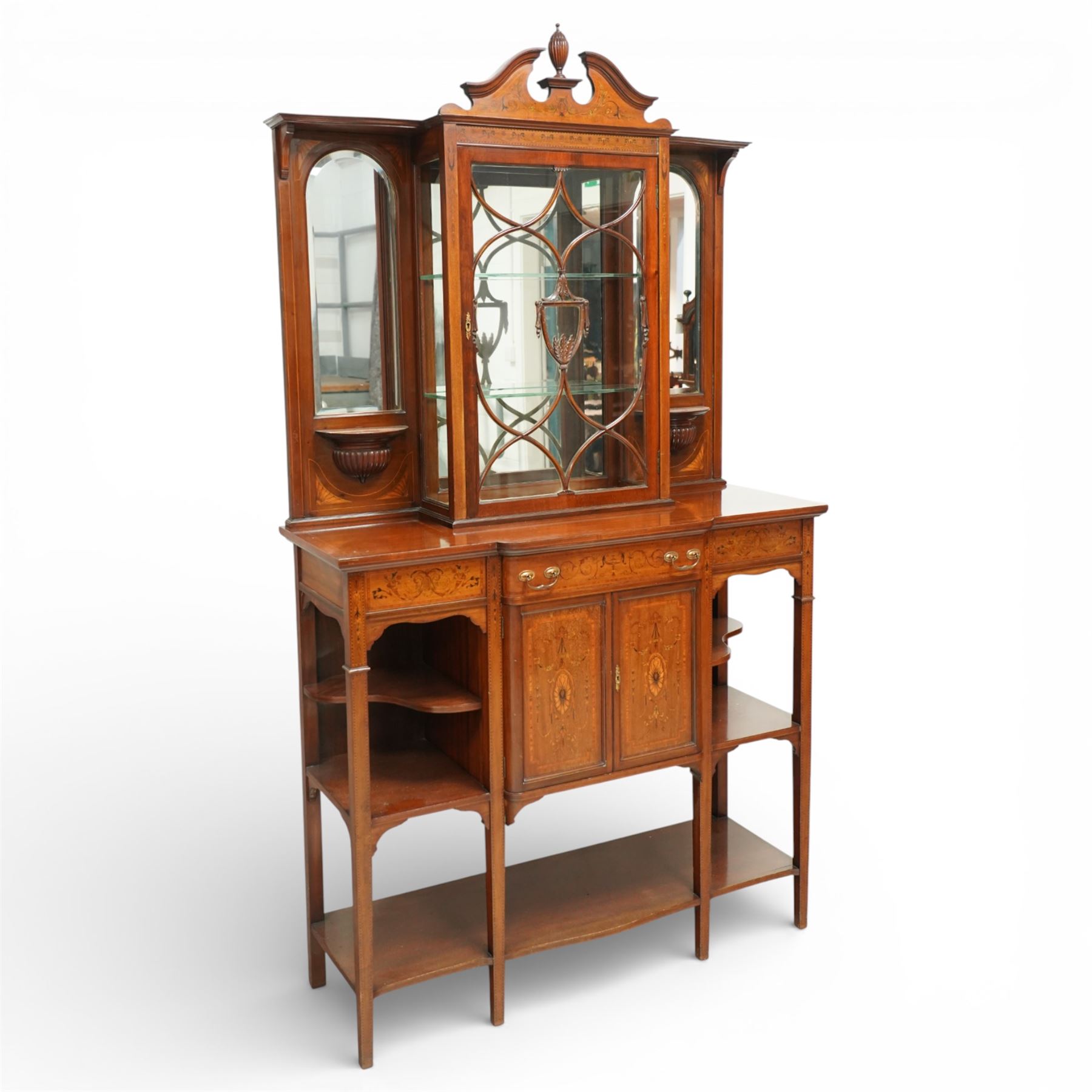Edwardian Sheraton Revival inlaid mahogany breakfront cabinet, the broken swan neck pediment with central finial inlaid with cartouche and scrolling floral festoons, the central cabinet fitted with astragal-style glazed door with central urn and swag motif, flanked by arched bevelled mirrors over a shelf and gadrooned bracket over paterae inlays, base fitted with marquetry frieze rawer over a panelled cupboard with intricate inlaid scrolling floral designs, flanked by open shelves, raised on square tapering herringbone inlaid supports united by undertier