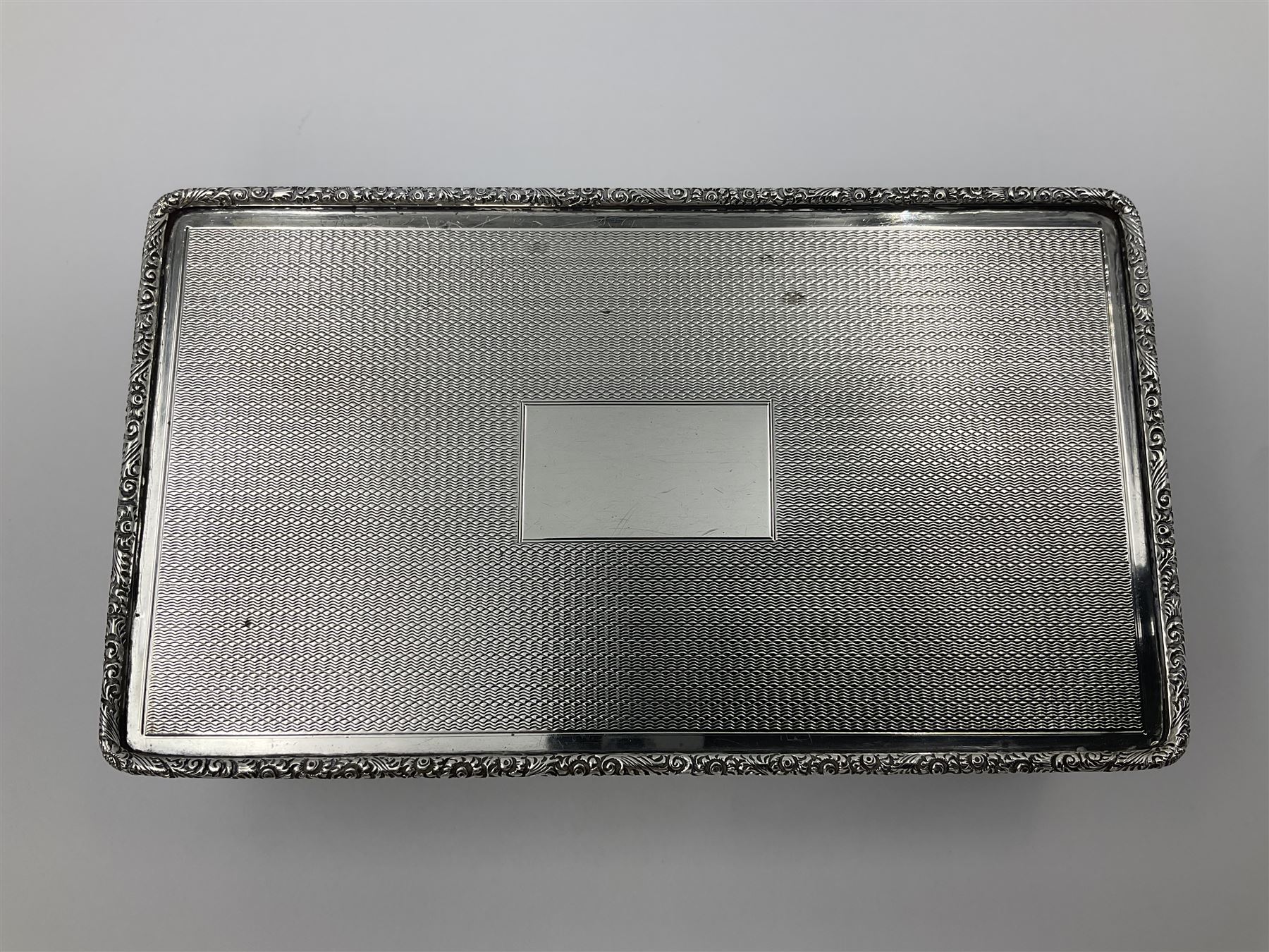 1920's silver table cigarette box, of rectangular form with engine turned decoration to the exterior, the hinged cover with central vacant rectangular panel and raised foliate rim, opening to reveal a gilt interior with later plush lining, hallmarked Charles Henry Dumenil, London 1923, H3.5cm L14.5cm D8.5cm