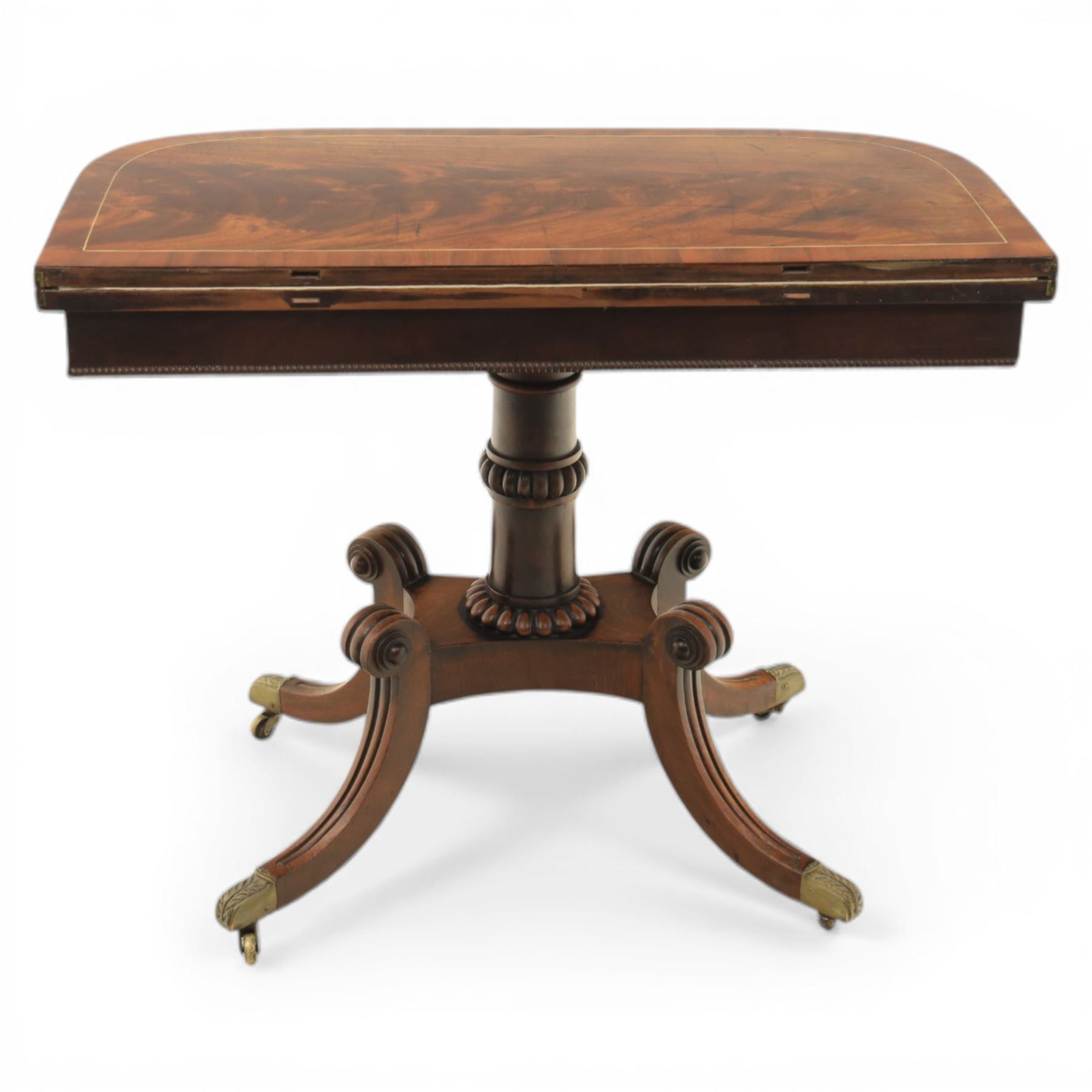 Early 19th century Regency mahogany fold over card table, the rounded rectangular top with brass line inlay enclosing green baize playing surface, crossbanded and strung exterior, with reeded edge above a gadrooned frieze, raised upon a turned column and four reeded sabre supports terminating in brass caps and castors