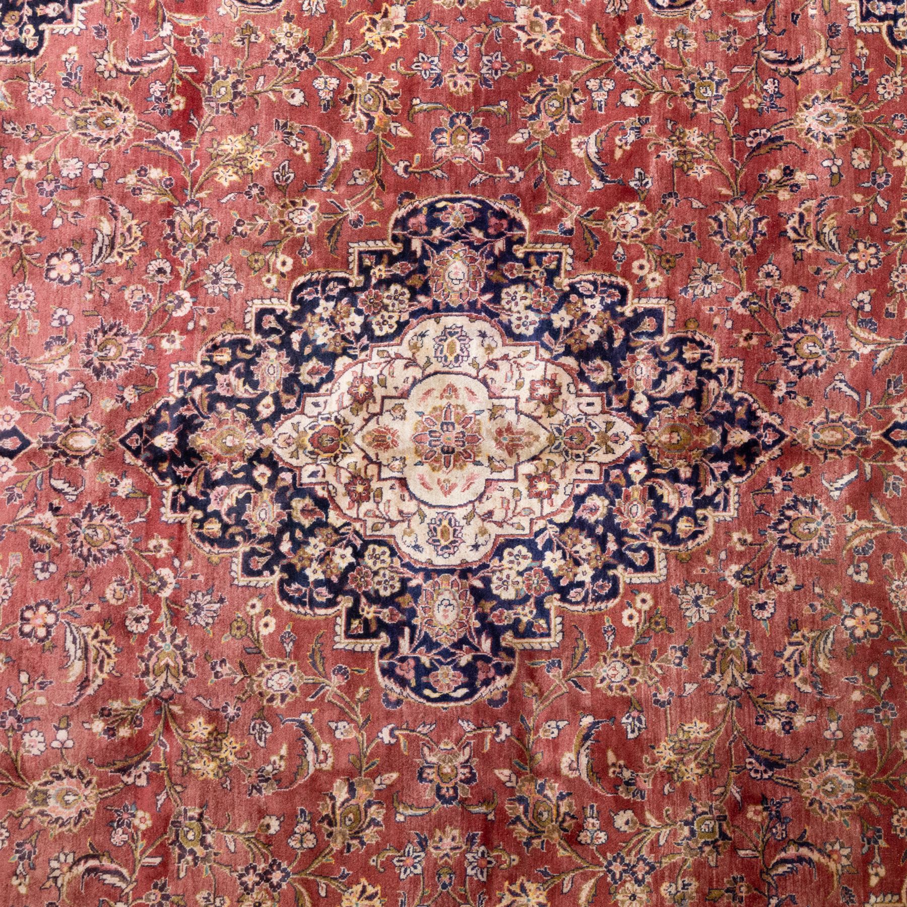 Large Central Persian Kashan hand-knotted carpet, madder red field with central cusped medallion on beige ground enclosed by indigo border with an intricate all-over arrangement of scrolling vines and floral motifs within conforming spandrels and a indigo ground main border ornamented with floral designs, enclosed by guard stripes and cotton fringe ends 