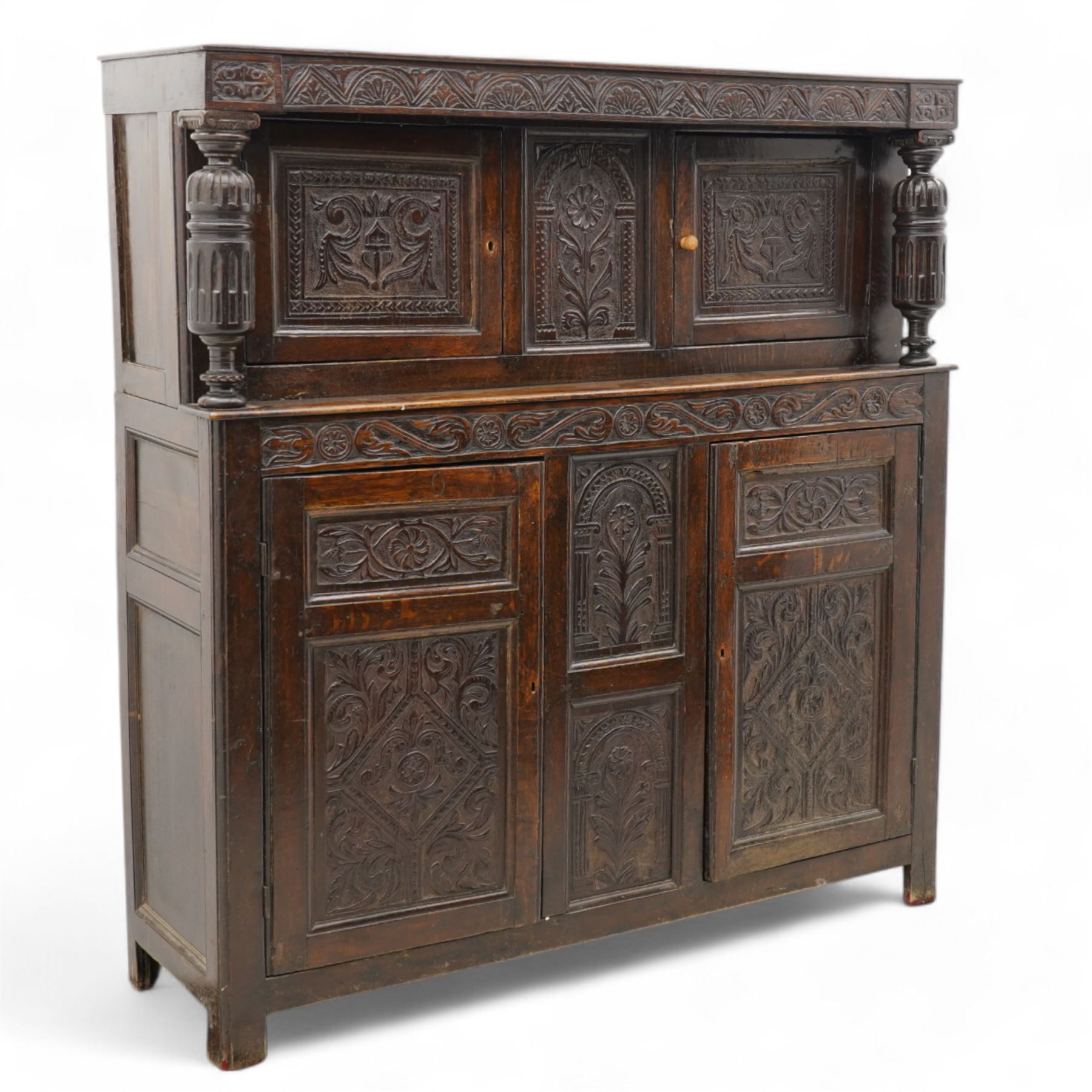 William & Mary design oak court cupboard, lunette carved frieze rail on turned and stop-fluted columns, the upper section fitted with two panelled doors carved with scrolling foliage and shield within a chip-carved border, central panel carved with arcade surrounding flower, foliate S-scroll lower frieze rail over two further cupboards and central panels, each carved with lozenges and curling foliage, moulded and pegged frame, terminating to stile supports 