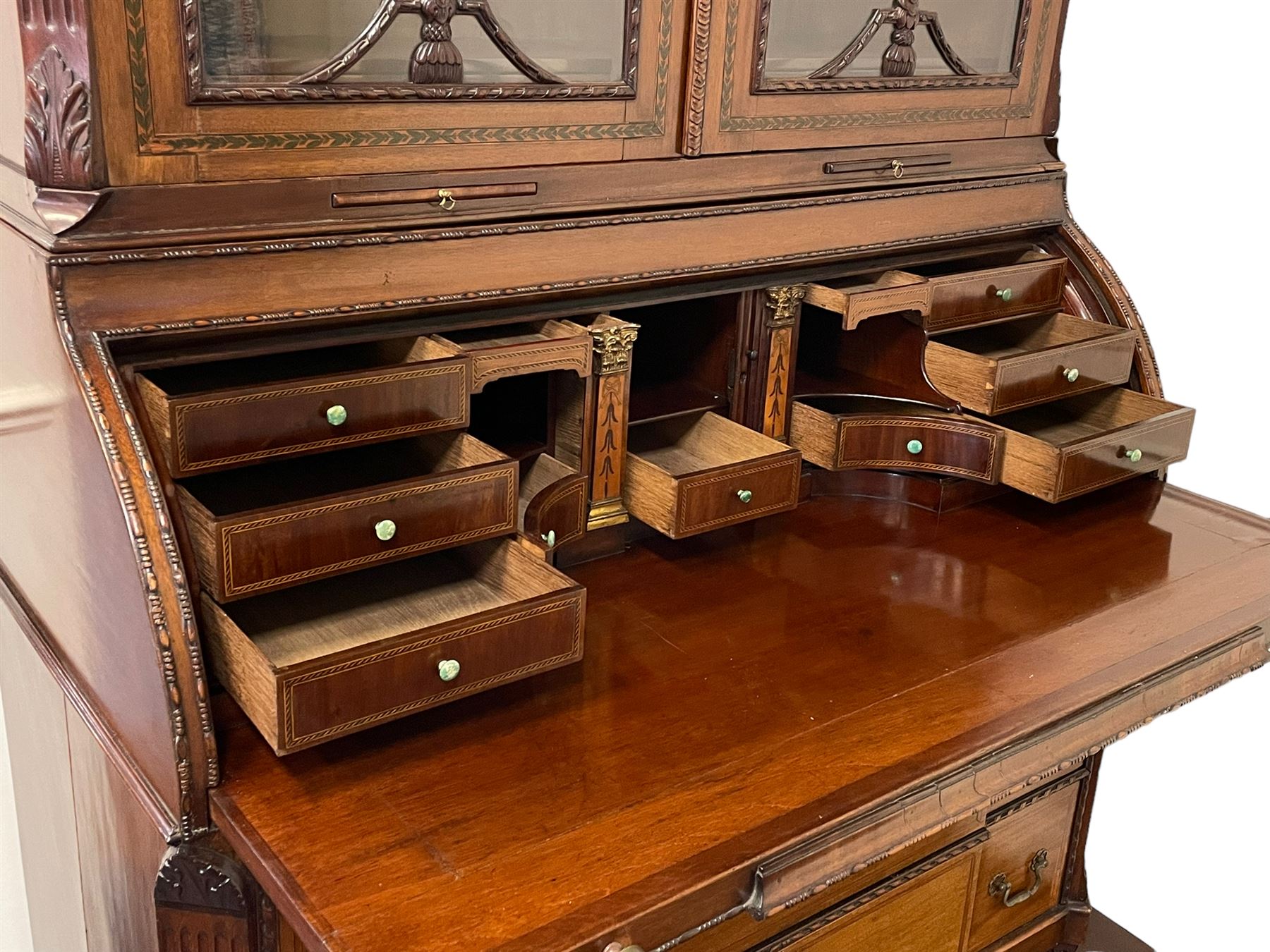 Large and impressive late 19th century Irish mahogany secretaire bookcase, moulded sloped arch dentil pediment decorated with central carved mask surrounded by linen festoons and trailing bellflowers, canted and fluted uprights with carved acanthus leaf upper and lower brackets, enclosed by two astragal glazed doors in the form of trophies with twist ribbon carved mouldings, inlaid with fanned spandrels and trailing leaf painted satinwood band, cylinder roll top inlaid with central fan motif and fanned spandrels, pull-out action revealing stepped fitted interior, central tambour roll cupboard flanked by two Composite pilasters and a combination of drawers and pigeon holes, fitted with three break-front drawers, matched canted and fluted corners with acanthus leaf carvings, on square tapering and fluted spade feet