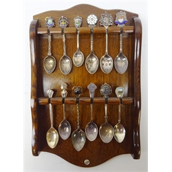  Collection of commemorative & crested silver and enamel teaspoons and one silver-plated, displayed on oak spoon rack   
