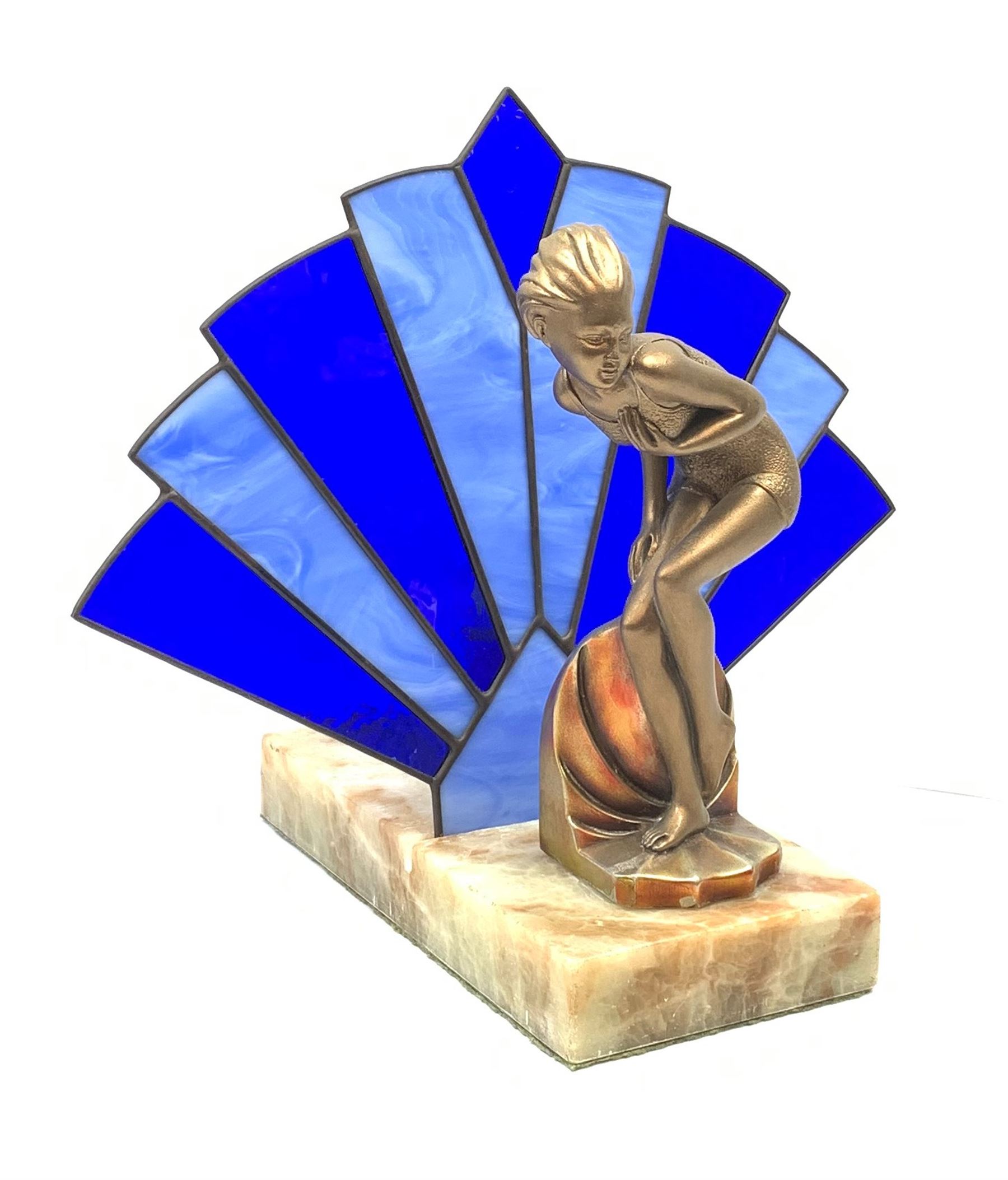 An Art Deco style table lamp, modelled in the form of a bronzed female figure before a leaded blue glass fan shaped shade, upon rectangular onyx base, overall H32cm base L28cm. 