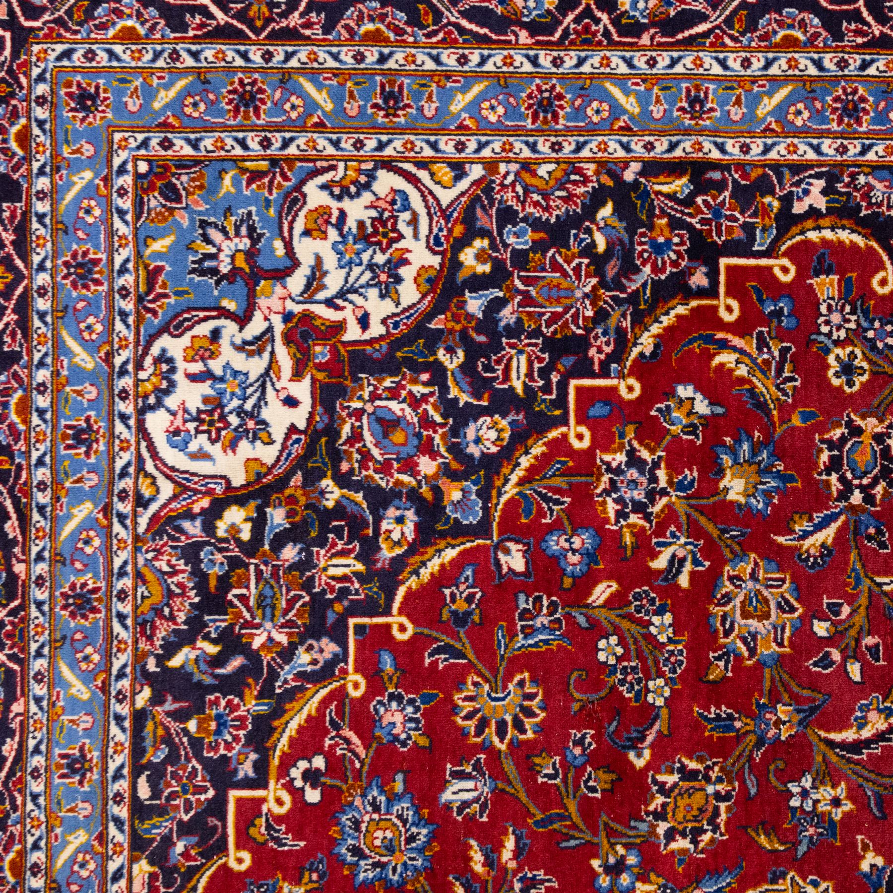 Central Persian Kashan hand-knotted carpet, rich crimson red field with a central cusped medallion with finely drawn floral sprays surrounded by dense scrolling vine work and palmettes, framed by a broad indigo ground border of flowering arabesques flanked by multiple guard stripes, in a palette of red, blue, ivory, sage green, and soft ochre tones, with cotton fringe ends    