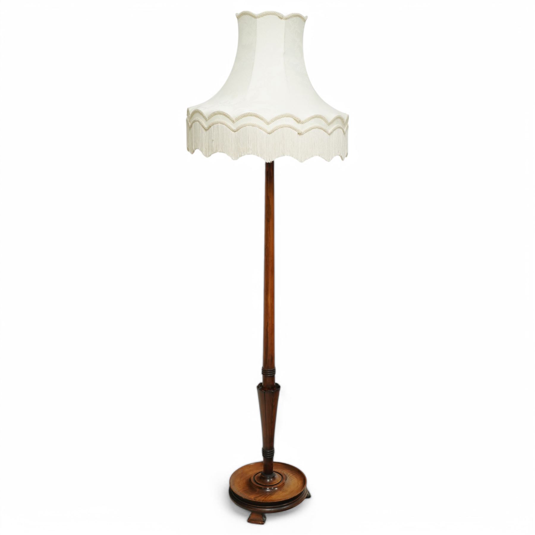 20th century walnut standard lamp, flared and faceted column over circular stepped base with shaped feet, with later scalloped cream fabric shade and fringe trim