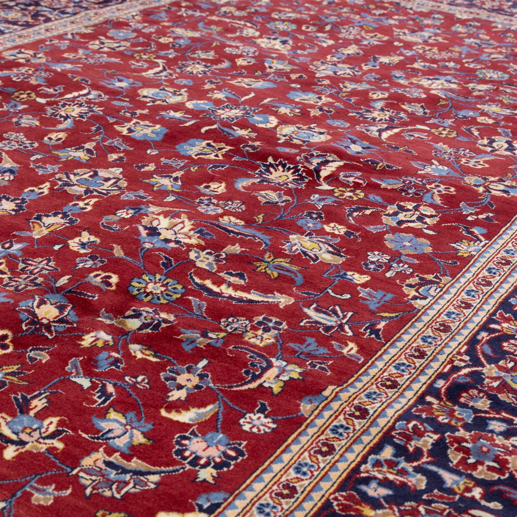 Central Persian Kashan hand-knotted rug, crimson field, decorated with an all-over floral and foliate design incorporating stylized palmettes, rosettes, and scrolling vine work, enclosed by a dark navy blue main border ornamented with flowering branches and palmettes flanked by multiple narrow guard borders with repeating floral motifs, with cotton fringe ends 