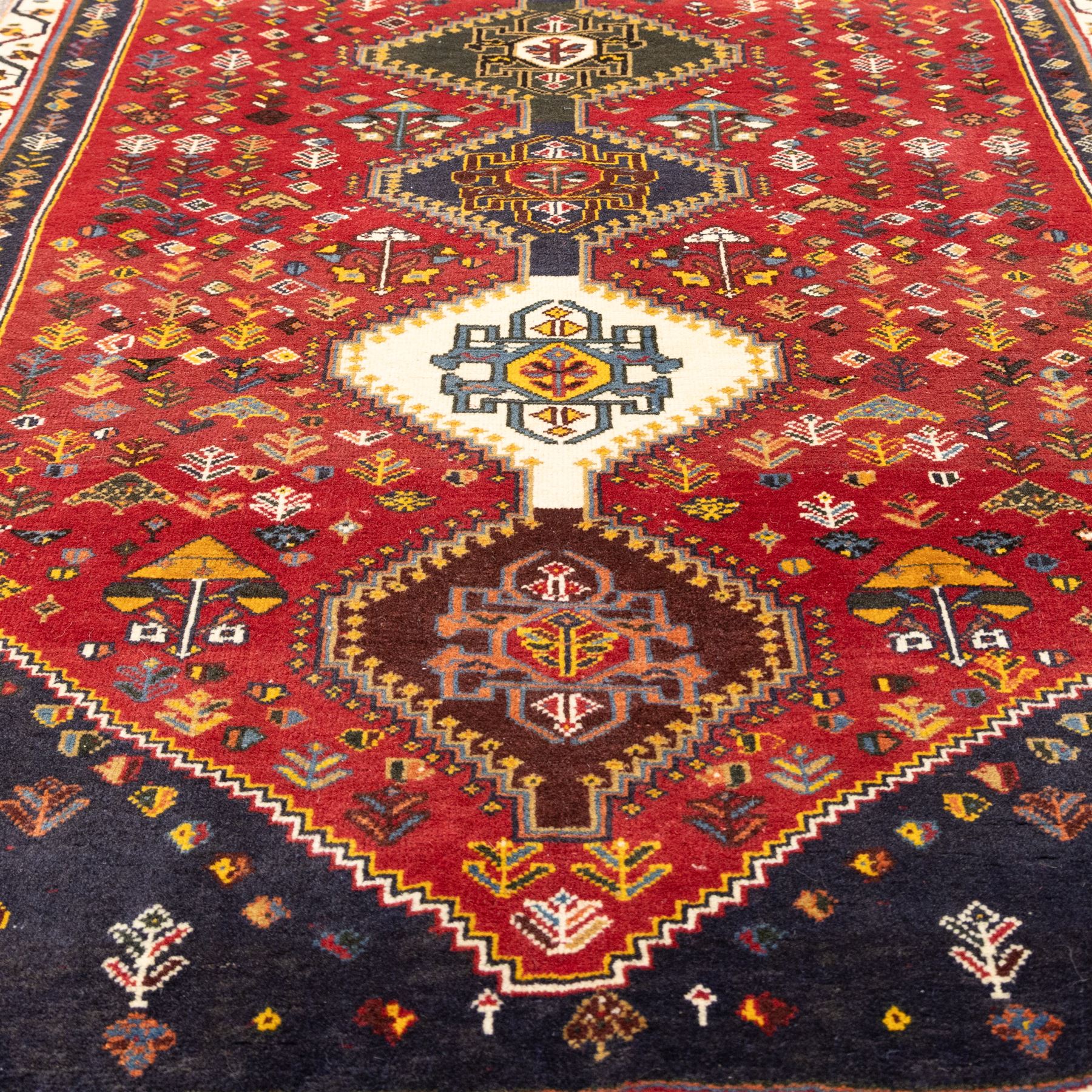 South West Persian hand-knotted Qashgai runner, madder-red field with a central column of linked diamond medallions in tones of indigo, ochre, ivory, and green, surrounded by stylized floral and boteh motifs within a cream border of angular vines and serrated devices, with cotton fringe ends   