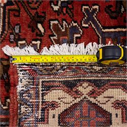 North West Persian Heriz hand-knotted carpet, madder red field with central stepped geometric medallion with radiating angular motifs and serrated outlines, and stylized floral and tribal devices framed by a broad border of repeating angular palmettes and rosettes flanked by multiple guard stripes, with cotton fringe ends 