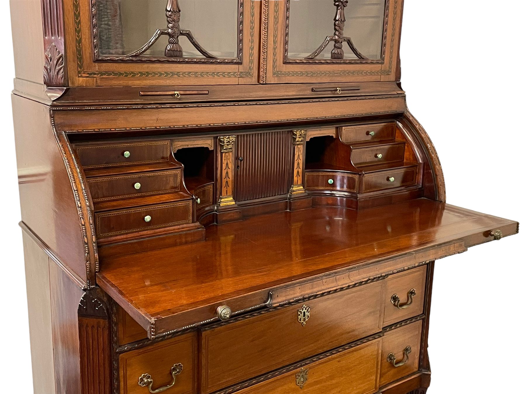 Large and impressive late 19th century Irish mahogany secretaire bookcase, moulded sloped arch dentil pediment decorated with central carved mask surrounded by linen festoons and trailing bellflowers, canted and fluted uprights with carved acanthus leaf upper and lower brackets, enclosed by two astragal glazed doors in the form of trophies with twist ribbon carved mouldings, inlaid with fanned spandrels and trailing leaf painted satinwood band, cylinder roll top inlaid with central fan motif and fanned spandrels, pull-out action revealing stepped fitted interior, central tambour roll cupboard flanked by two Composite pilasters and a combination of drawers and pigeon holes, fitted with three break-front drawers, matched canted and fluted corners with acanthus leaf carvings, on square tapering and fluted spade feet