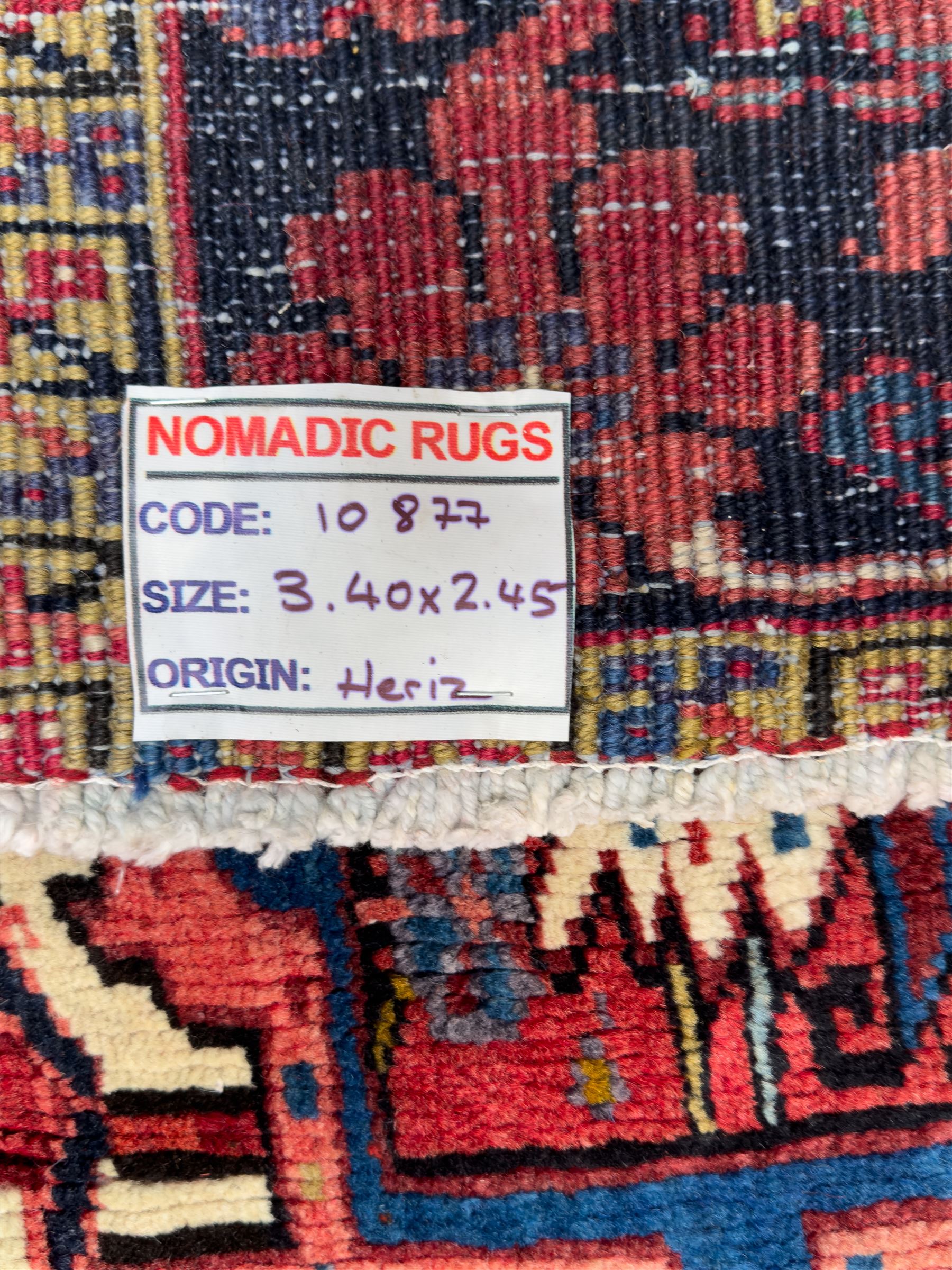 Northwestern Persian Heriz hand-knotted carpet, red madder field with a bold stepped medallion in indigo, ivory, and terracotta, flanked by angular floral motifs and stylized leaves, the spandrels set within a dark indigo border of rosettes and angular vine motifs between narrow guard stripes, 340cm x 245cm 