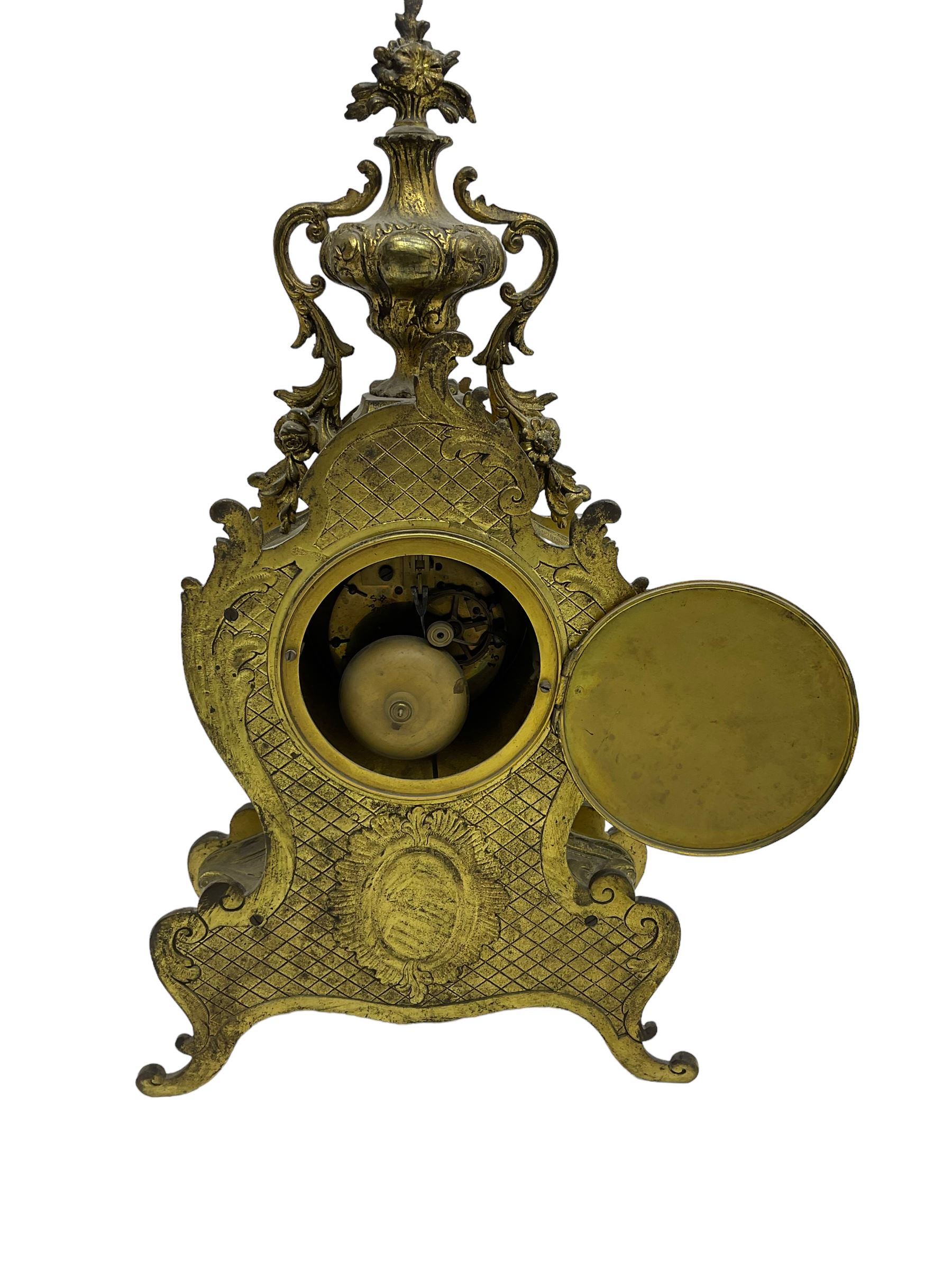 French - late 19th century 8-day gilt brass mantle clock surmounted by an urn, with a waisted case and baroque decoration raised on four splayed feet, with a glazed bezel, enamel dial with Roman numerals, five minute Arabic's and gothic steel hands, count wheel striking movement, striking the hours and half-hours on a bell. With pendulum.