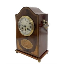 Edwardian- 8-day mantle clock in a mahogany case with a serpentine top and contrasting inlayed panels in satinwood and boxwood to the front, on a rectangular plinth raised on ball feet, silvered dial with Roman numerals and steel spade hands, twin train Parisian rack striking movement, sounding the hours and half hours on a coiled gong. With pendulum.